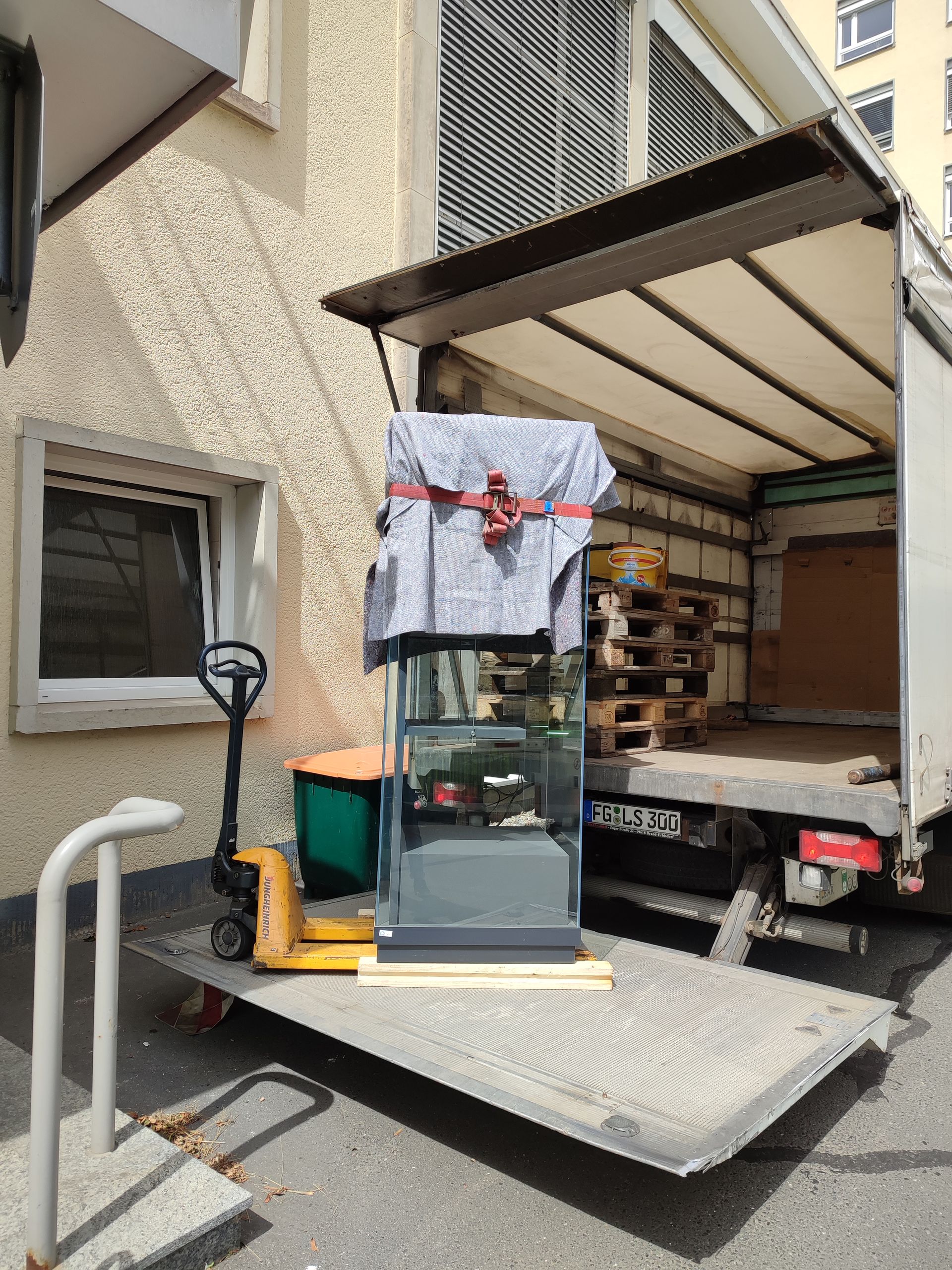 Ein großes, mit Stoff umwickeltes Aquarium wird neben einem Gebäude mithilfe eines Hebezeugs auf einen Lastwagen verladen.