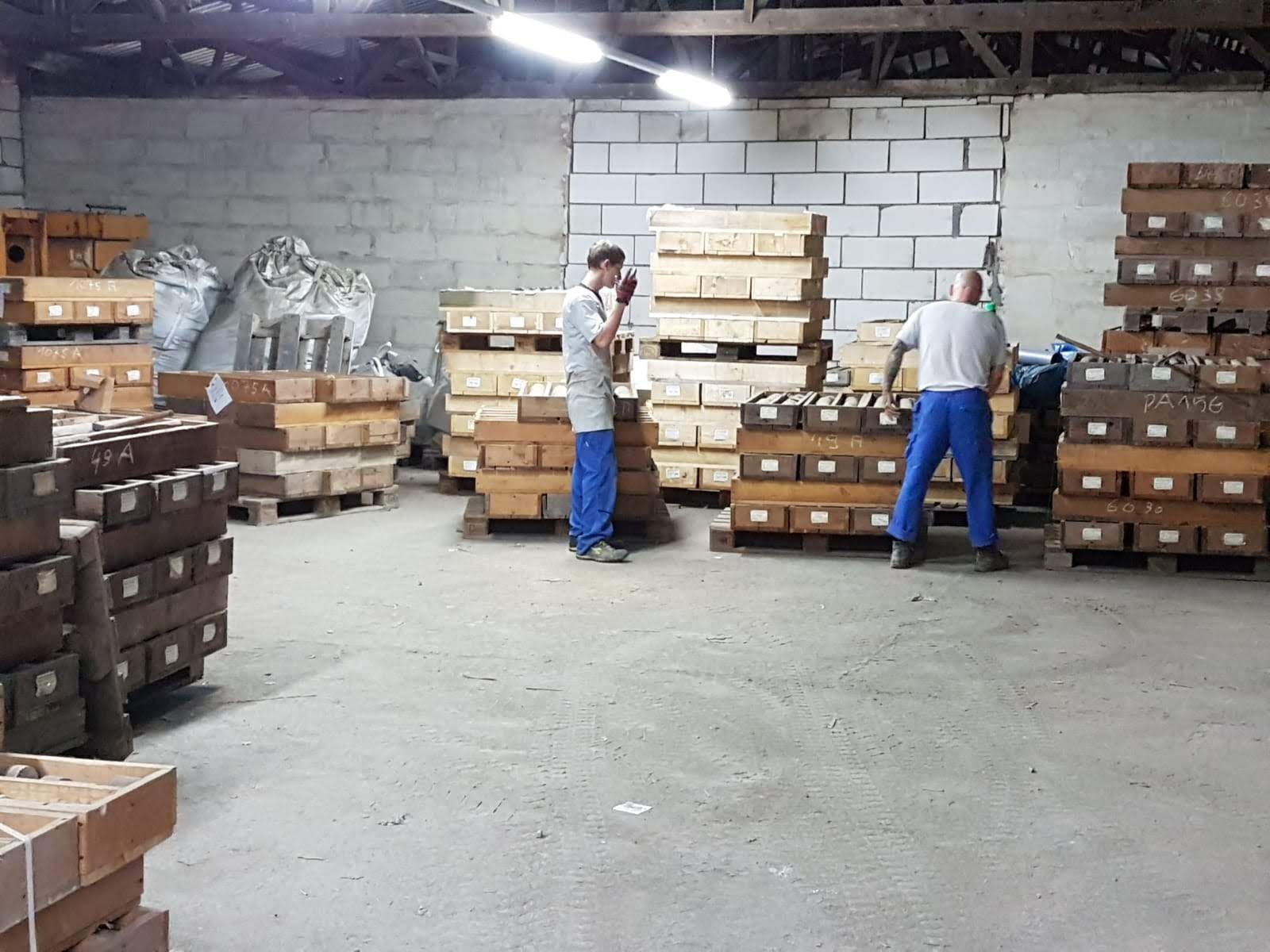 Zwei Arbeiter in einem Lagerhaus, umgeben von gestapelten, rechteckigen Gegenständen.