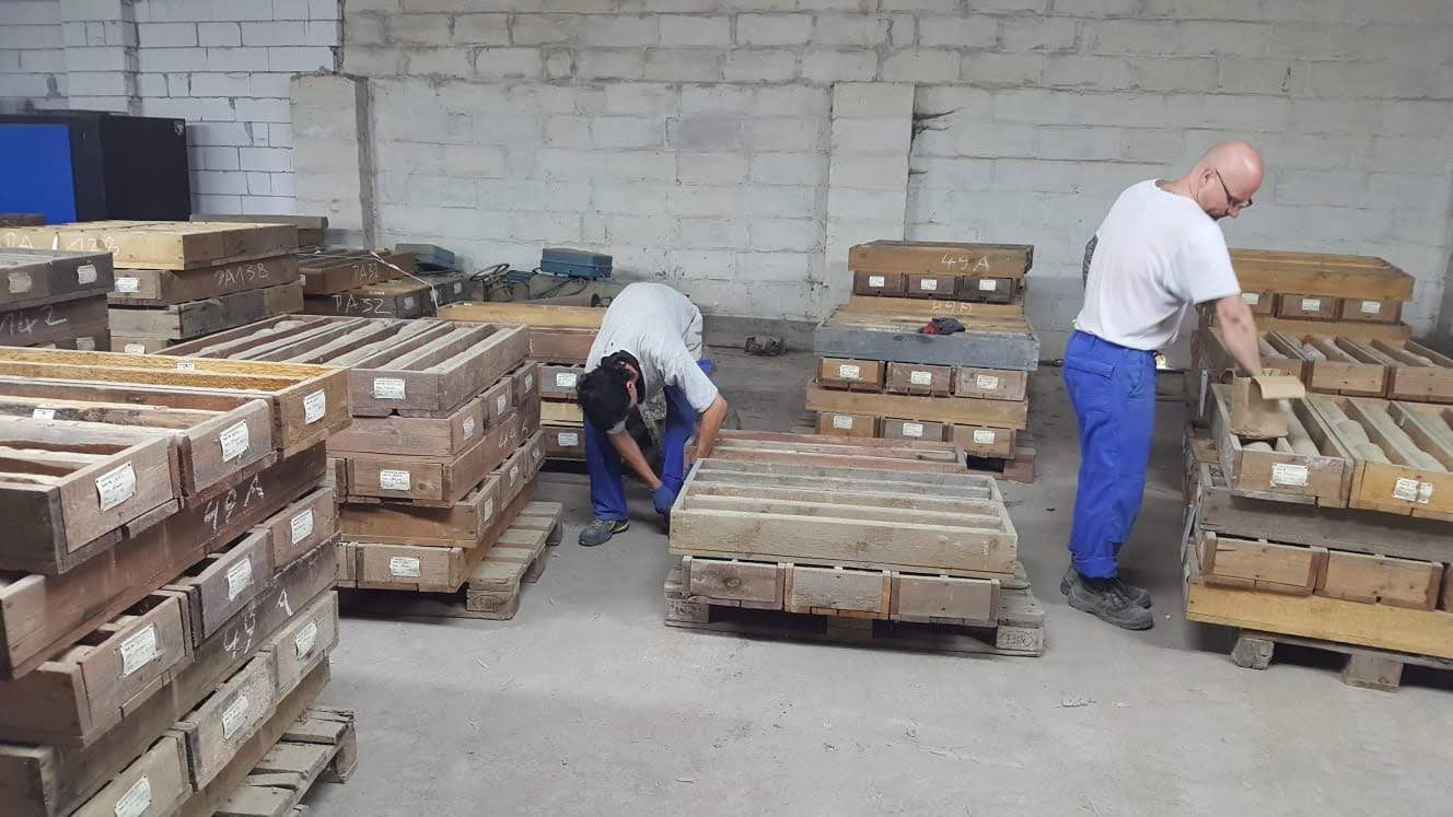 Zwei Personen stapeln in einem Lagerhaus Holzpaletten mit rechteckigen Blöcken.