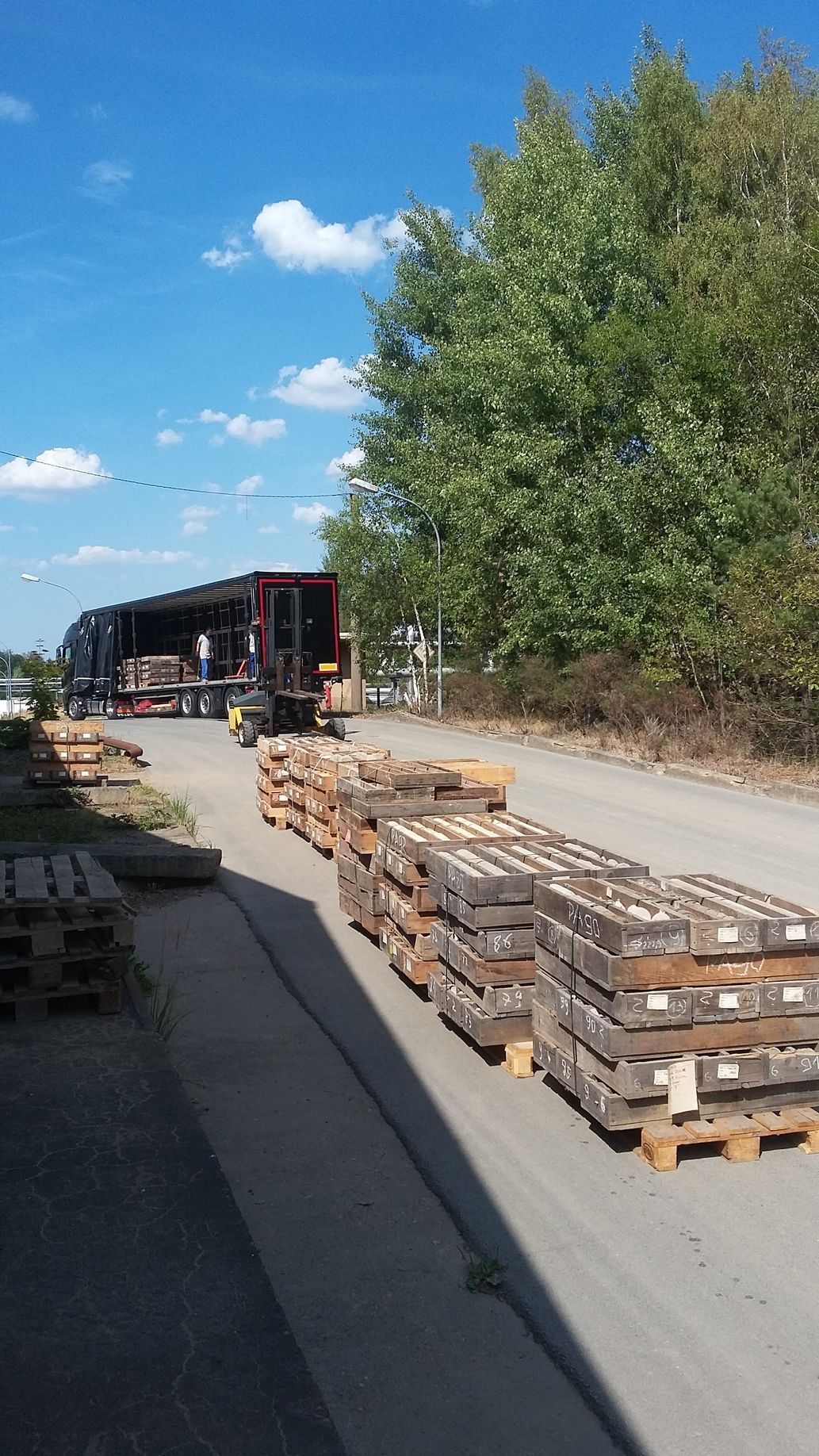 Auf einer Straße sind Paletten gestapelt, im Hintergrund wird unter blauem Himmel ein LKW beladen.