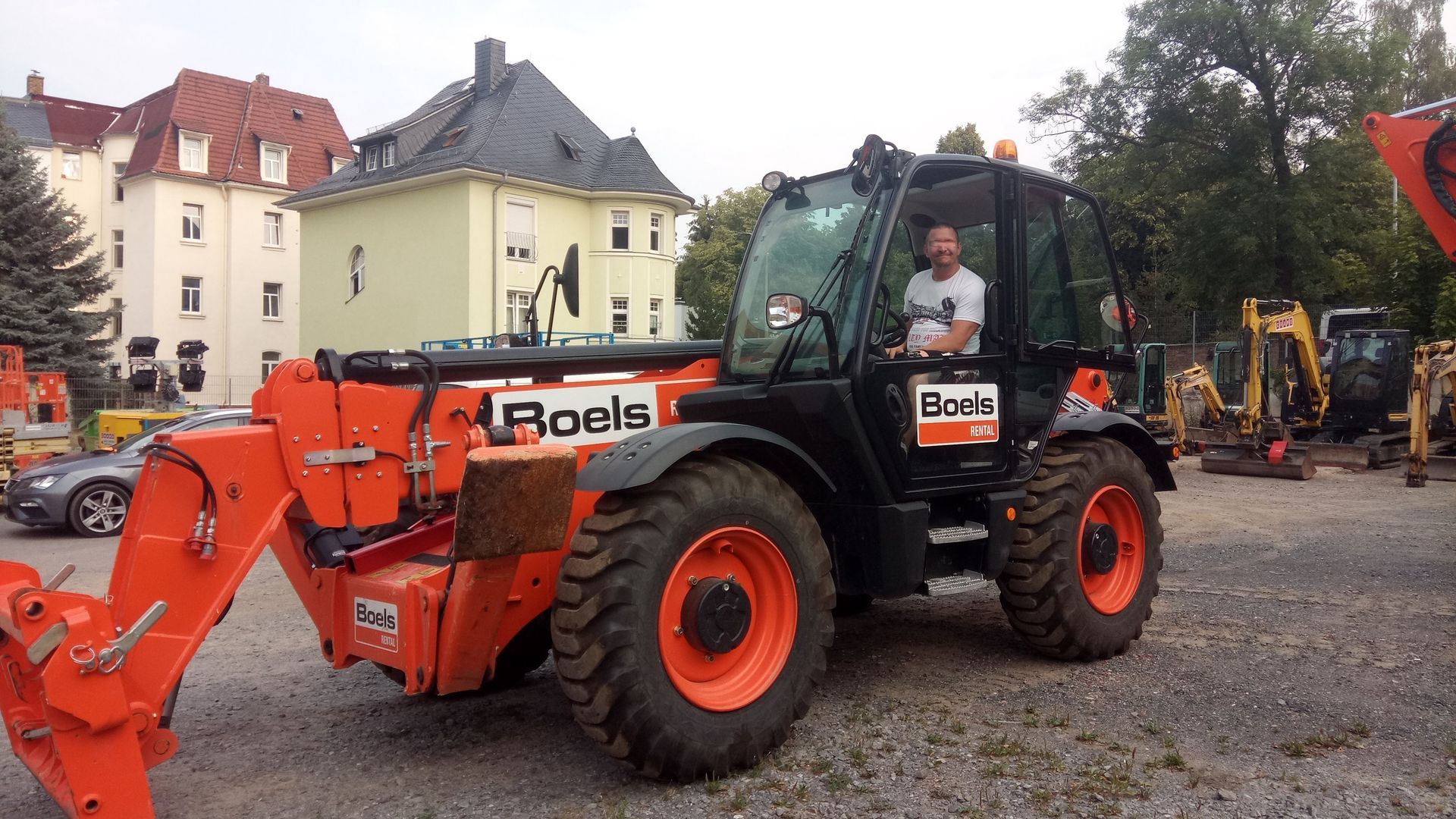 Mann in orange-schwarzem Boels-Teleskoplader auf einem Parkplatz, im Hintergrund Gebäude.
