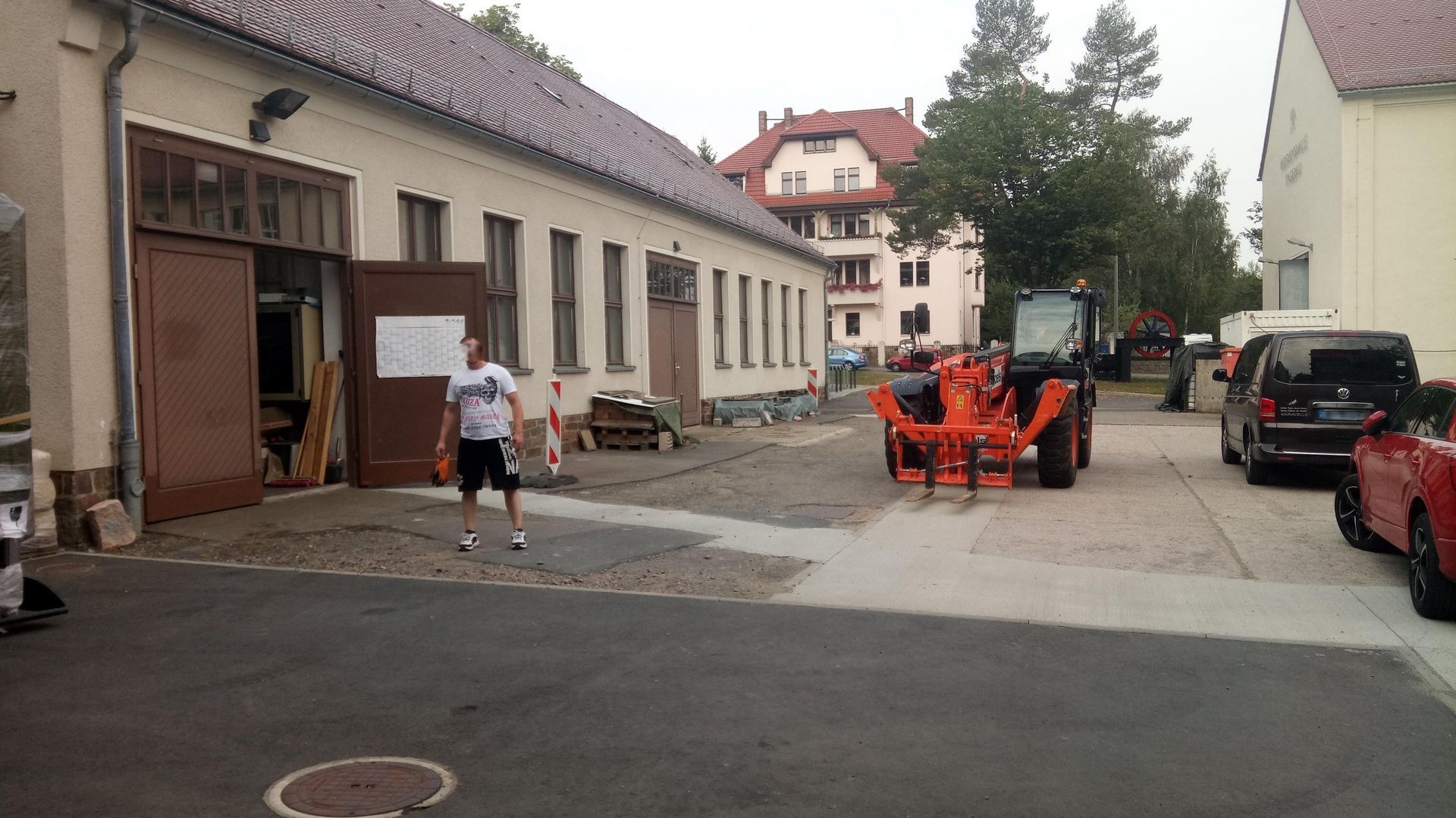 Ein Mann in Shorts und ein Traktor stehen vor einem Gebäude mit offenen Türen; in der Nähe sind Autos geparkt.