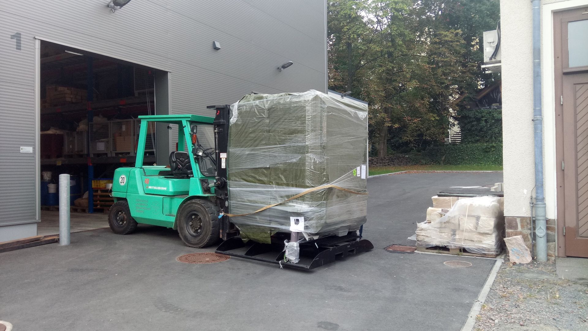 Ein grüner Gabelstapler verlädt ein großes, verpacktes Paket vor einem Lagerhaus.