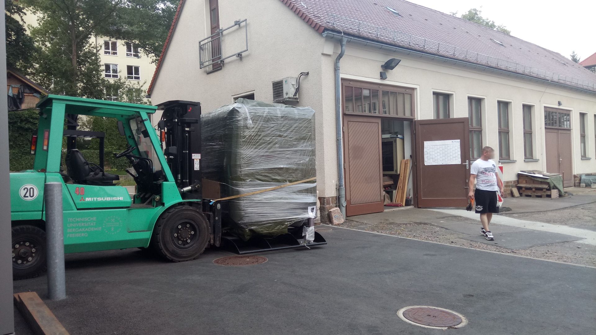 Ein grüner Gabelstapler transportiert eine große, verpackte Ladung in der Nähe eines Gebäudes; eine Person geht in der Nähe vorbei.