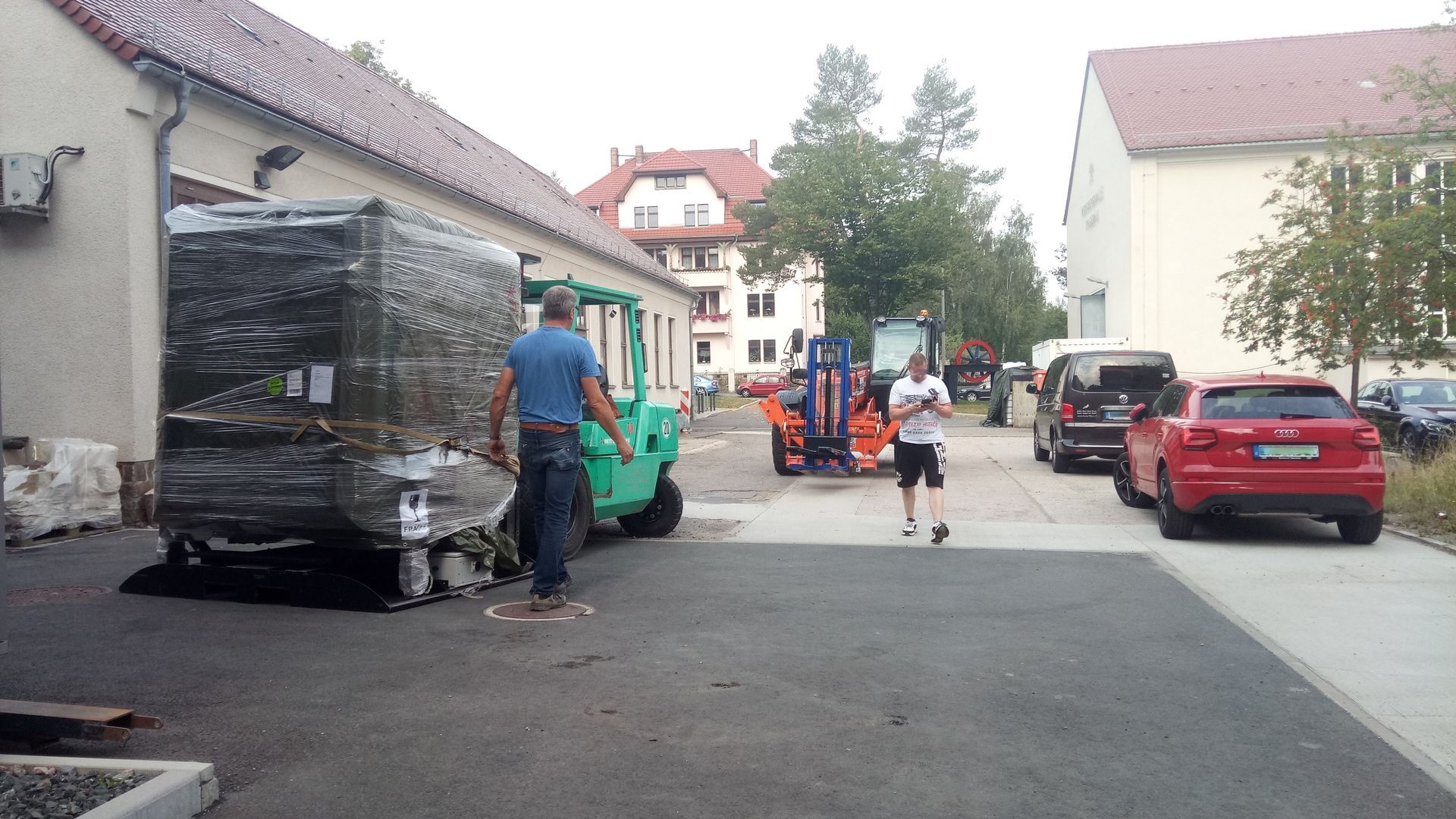 Ein Mann mit einem grünen Gabelstapler transportiert eine eingewickelte Palette in der Nähe von Gebäuden; weitere Fahrzeuge sind anwesend.