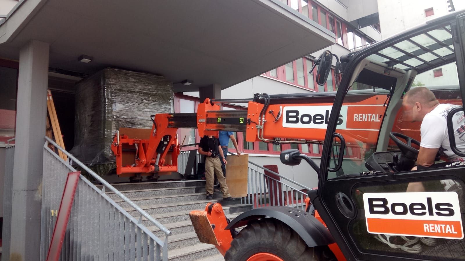 Ein Gabelstapler transportiert eine große Kiste eine Treppe hinauf in der Nähe eines Gebäudeeingangs; Arbeiter helfen dabei.