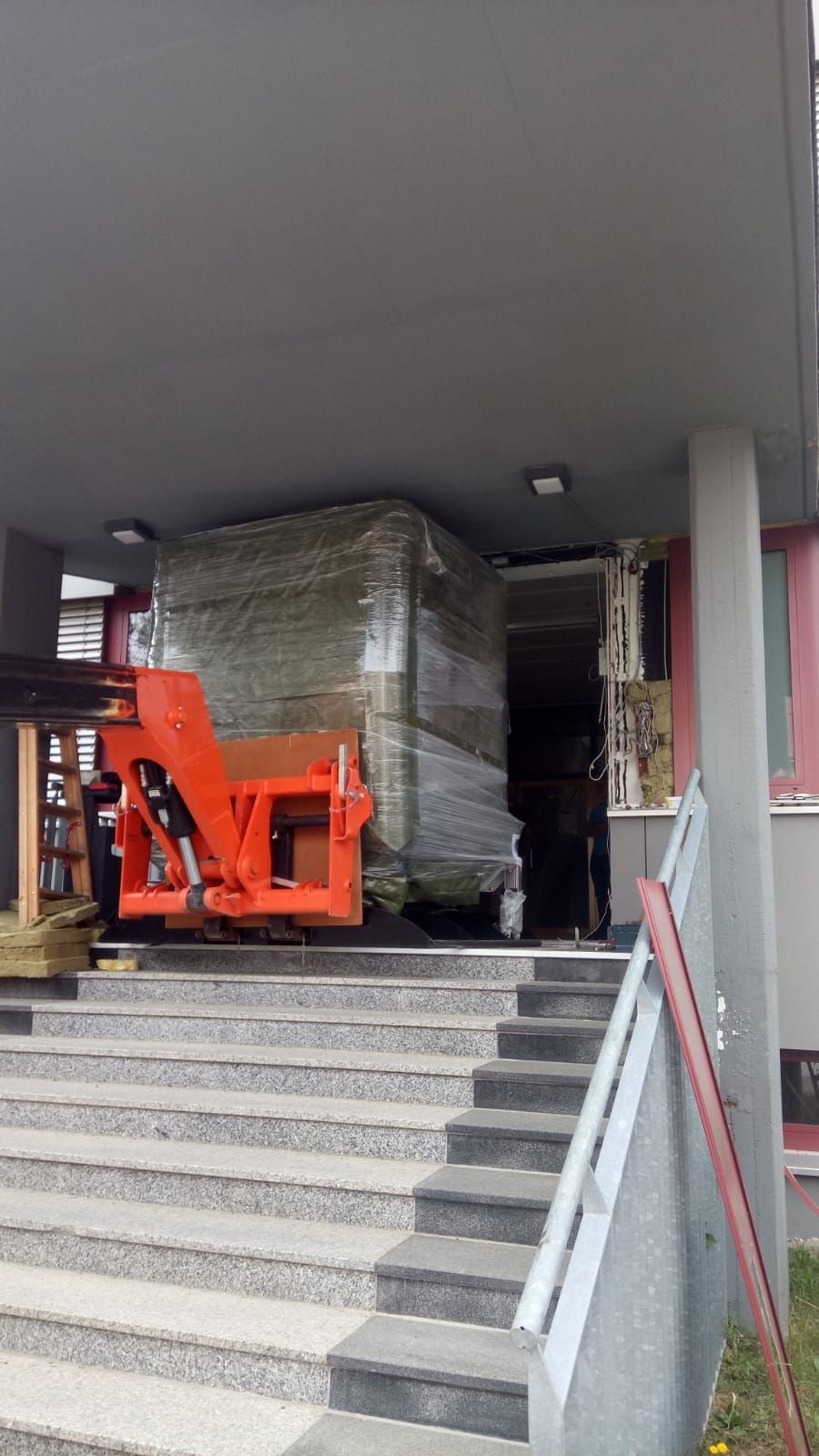 Ein orangefarbener Gabelstapler transportiert einen großen, verpackten Gegenstand die Treppe eines Gebäudes hinauf. Eine graue Treppe führt zu einem überdachten Eingang.