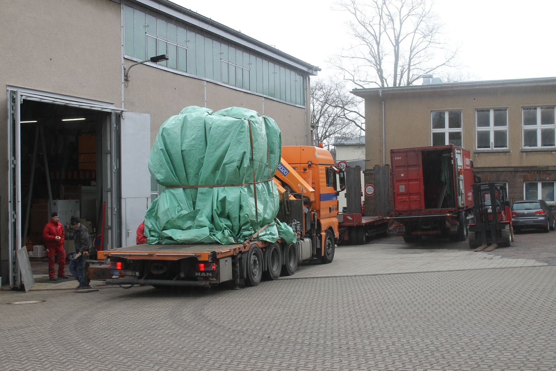 Ein großes, in eine grüne Plane gehülltes Objekt wird auf einem Pritschenwagen in der Nähe eines Gebäudes verladen.