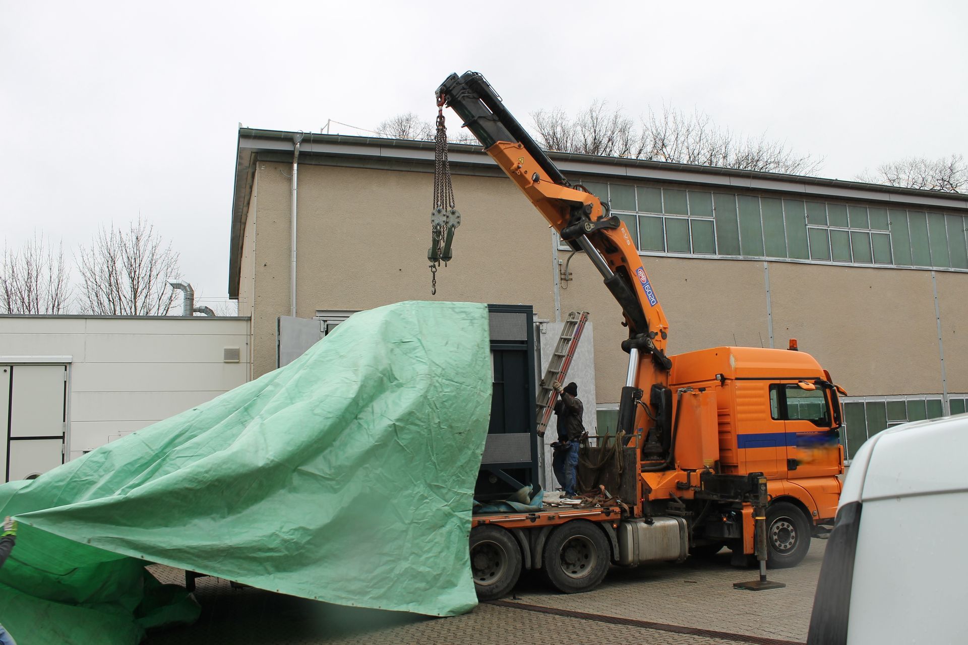 Ein orangefarbener Kran hebt einen abgedeckten Gegenstand von einem LKW in der Nähe eines Gebäudes.
