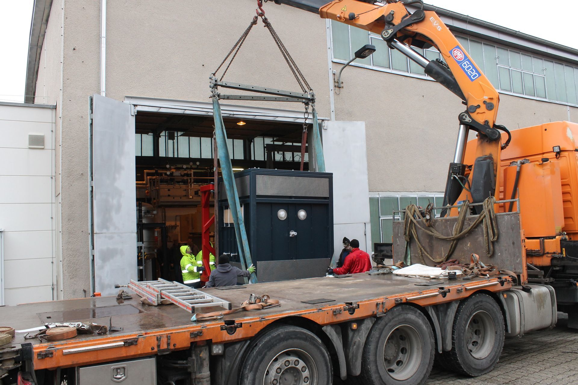 Ein Kran hebt Ausrüstung von einem Pritschenwagen in ein Gebäude; Arbeiter in Warnwesten und anderer Schutzausrüstung helfen dabei.
