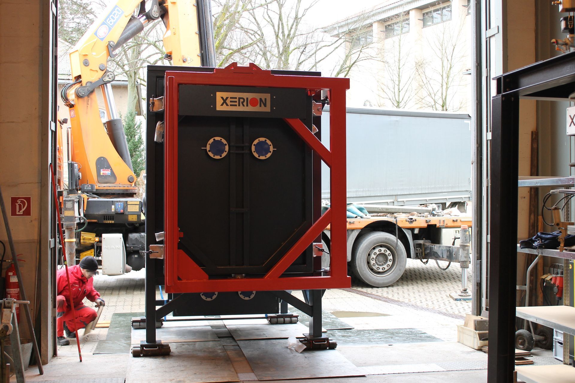 Eine große schwarze Industriehalle mit rotem Rahmen wird mit einem Kran in eine Garage gefahren.