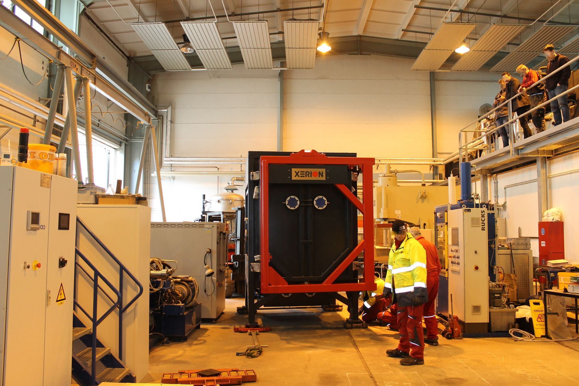 Fabrikinterieur mit Arbeitern in der Nähe einer großen schwarzen Maschine, die in einem roten Rahmen eingefasst ist; Menschen beobachten das Geschehen von einem Balkon aus.