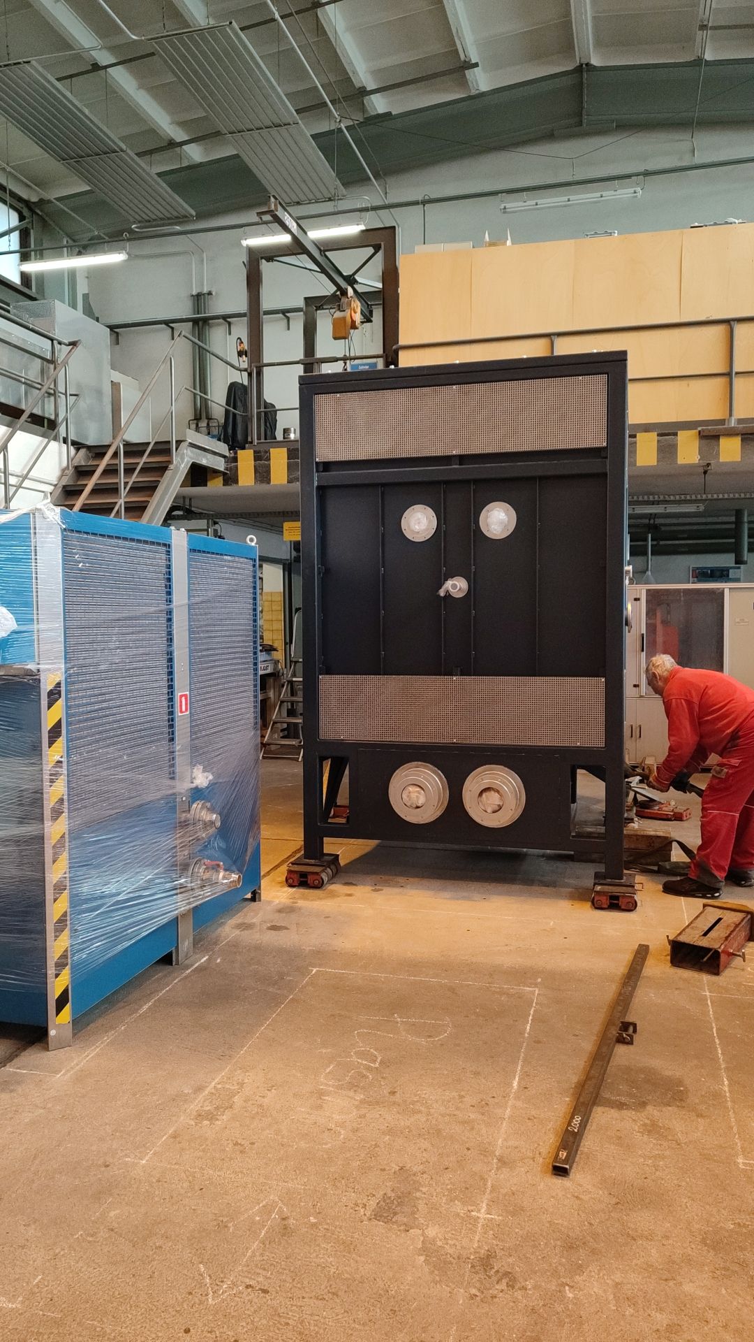 Schwarze Industriemaschine in einer Fabrik. Ein Arbeiter in rotem Overall arbeitet an der Maschine.