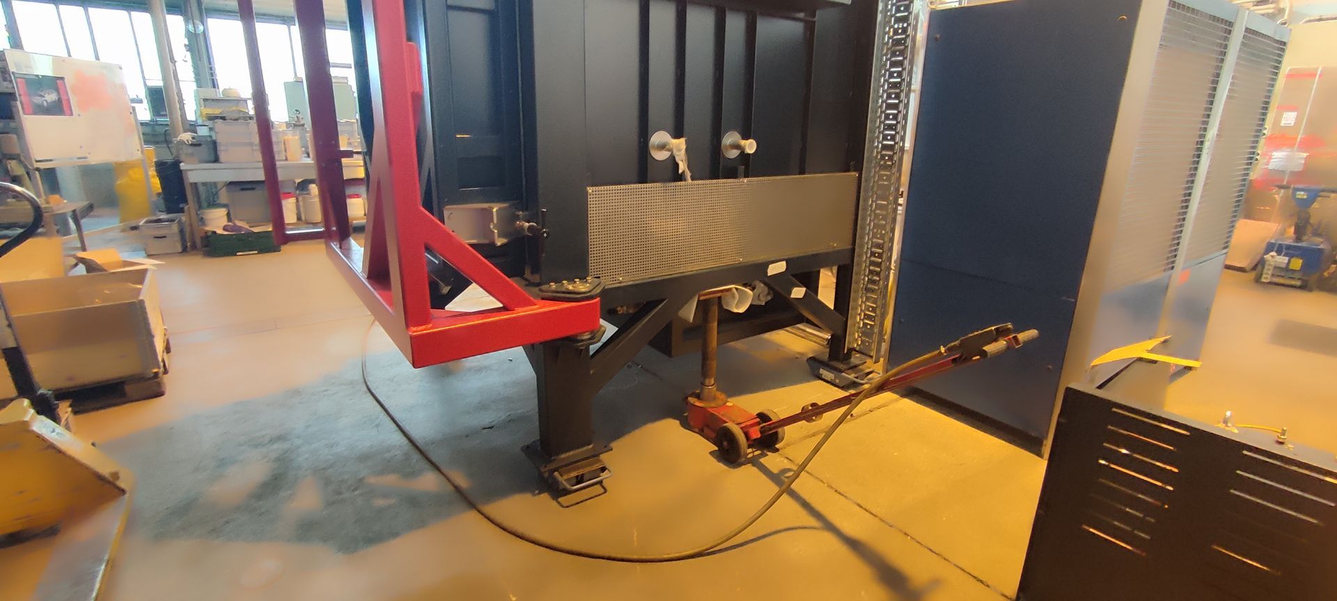 Eine große Industriemaschine mit rotem Metallrahmen und schwarz-blauen Verkleidungen. Auf dem Boden befindet sich ein Fußpedal.