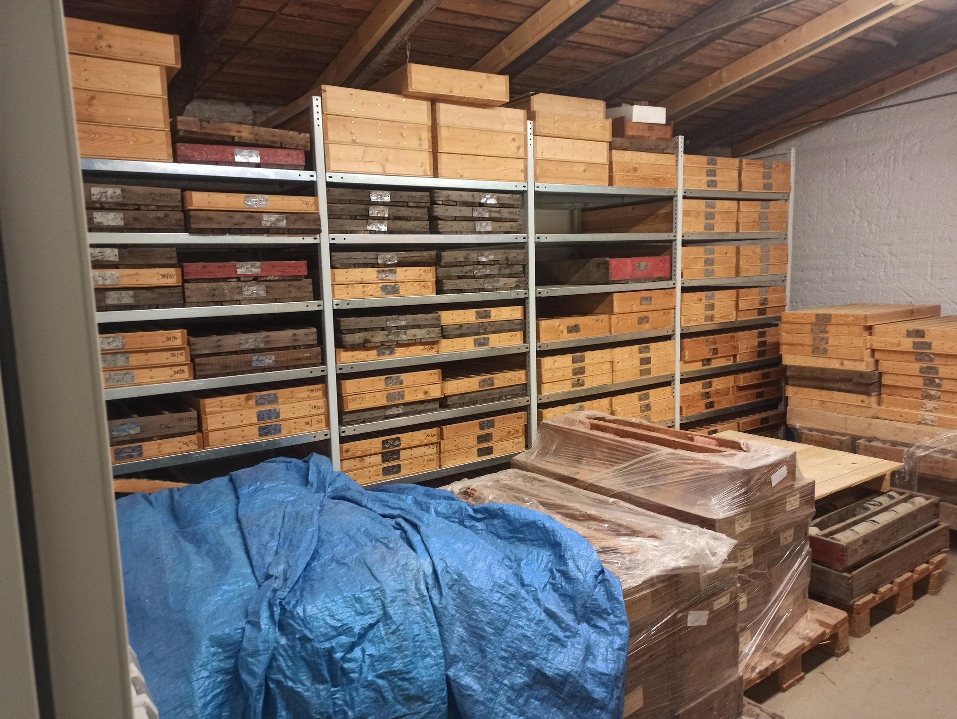 Im Lagerraum stehen Regale voller brauner Kisten und Behälter. Blaue Plane und Holzpaletten sind zu sehen.