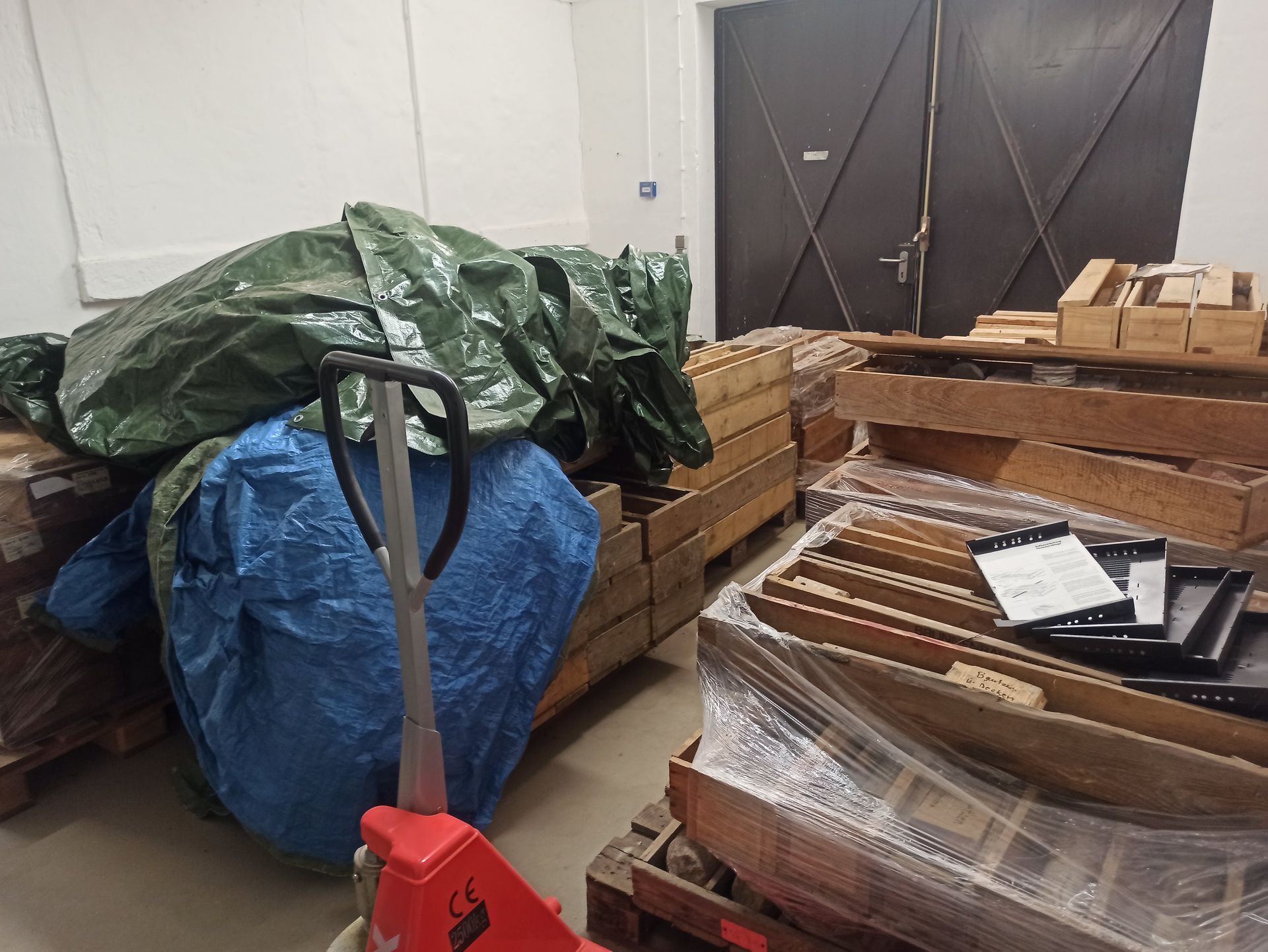 Innenansicht einer Lagerhalle mit Paletten voller Holzkisten, Planen und einem roten Hubwagen.