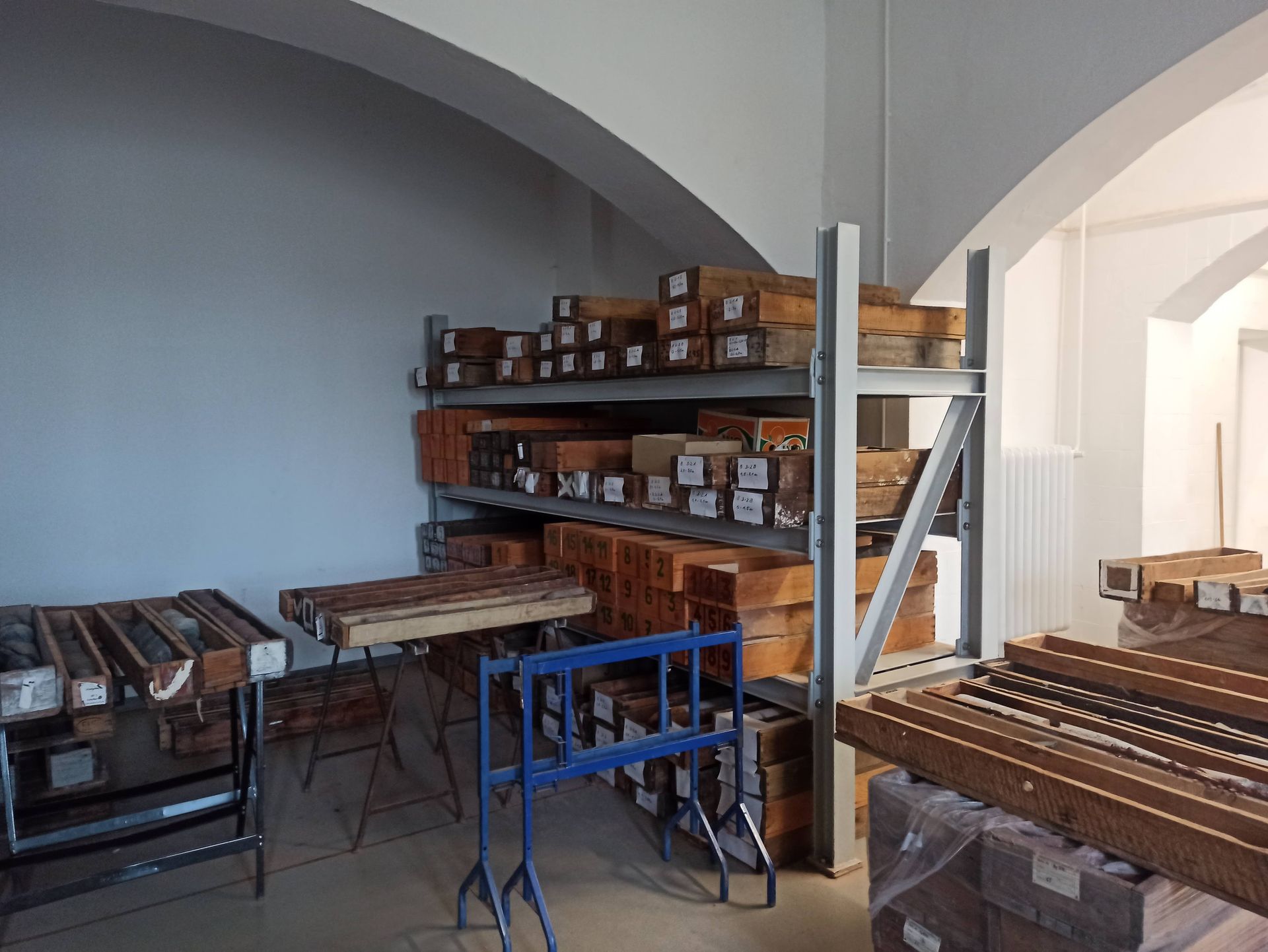 Holzbalken und Kisten auf Metallregalen in einem Abstellraum mit gewölbter Decke.