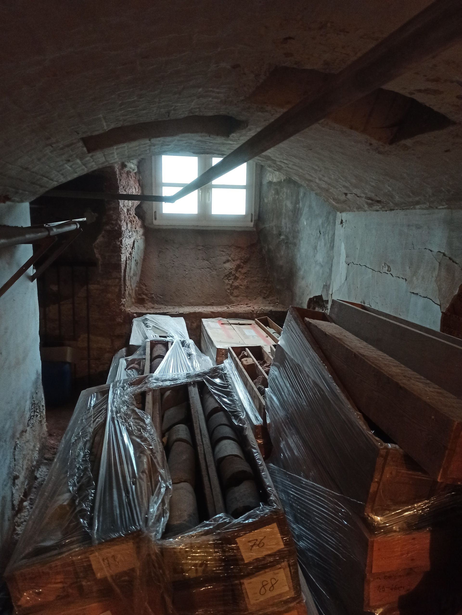 Innenansicht eines schwach beleuchteten Dachzimmers mit einem kleinen Fenster, verschiedenen eingewickelten Gegenständen und freiliegenden Ziegelwänden.