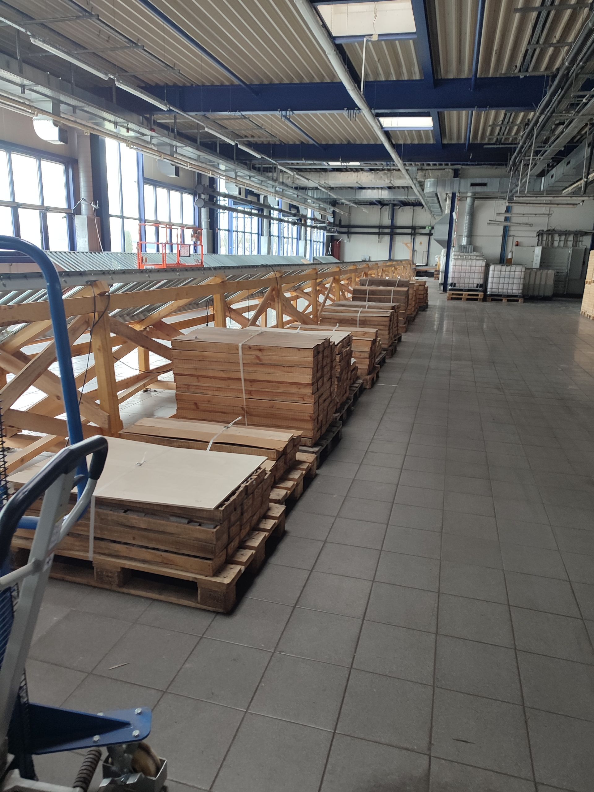 In einer Fabrik stehen gestapelte Holzpaneele auf Paletten neben einem Förderband.