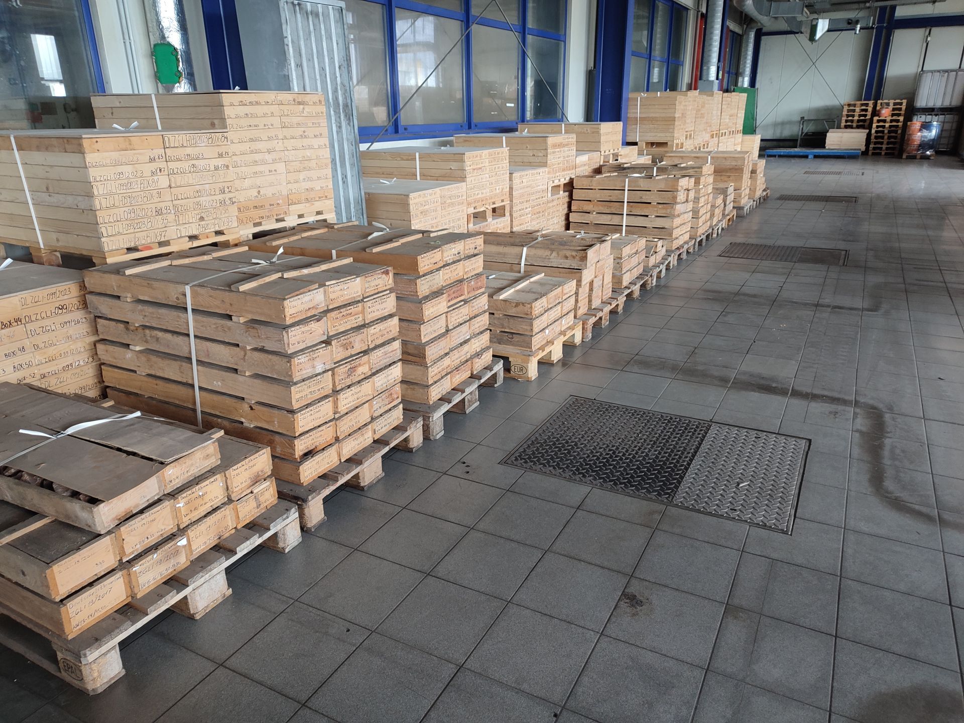 In einer Industrieumgebung sind Holzkisten auf Paletten gestapelt und in Reihen auf einem gefliesten Boden angeordnet.