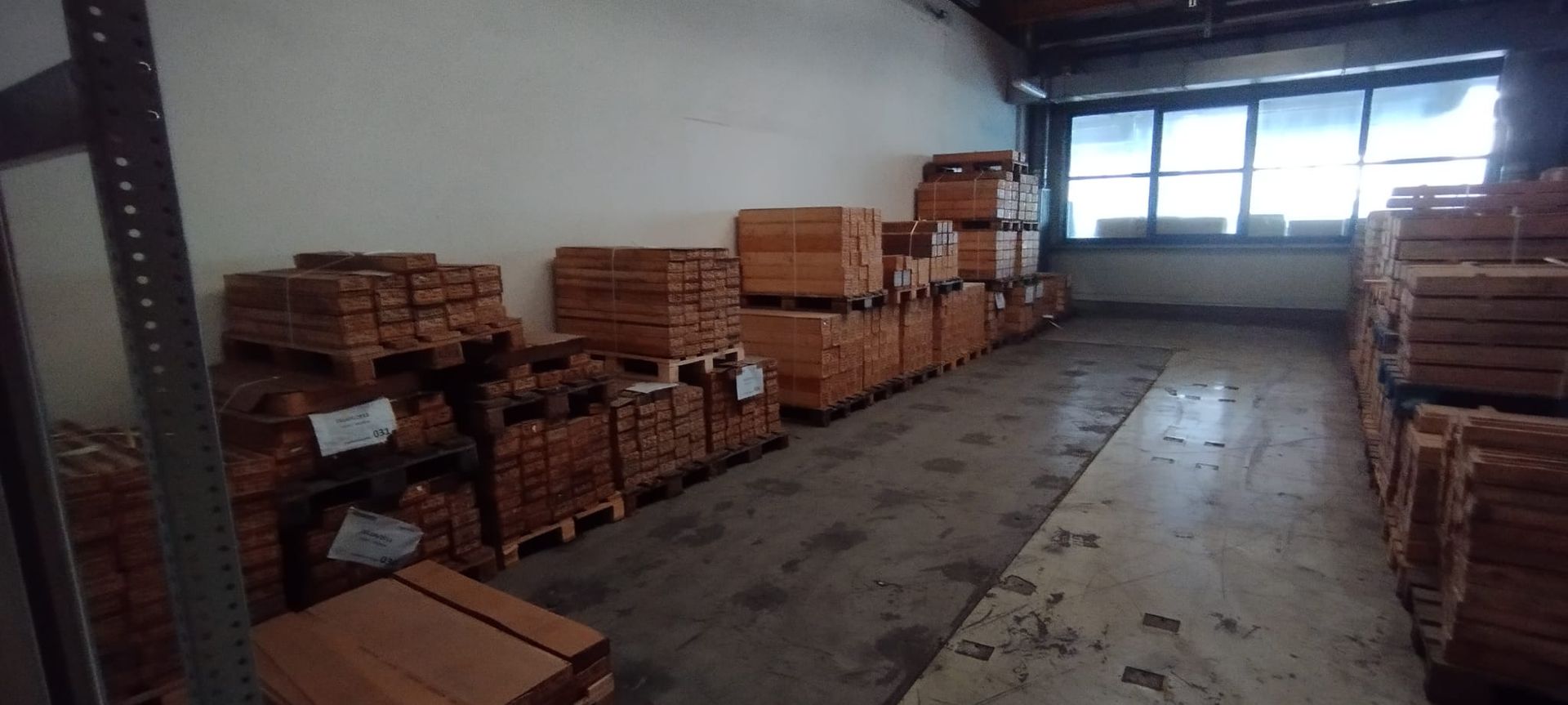 Innenansicht einer Lagerhalle mit gestapelten Holzplanken, Betonboden und Fenstern an der Rückwand.
