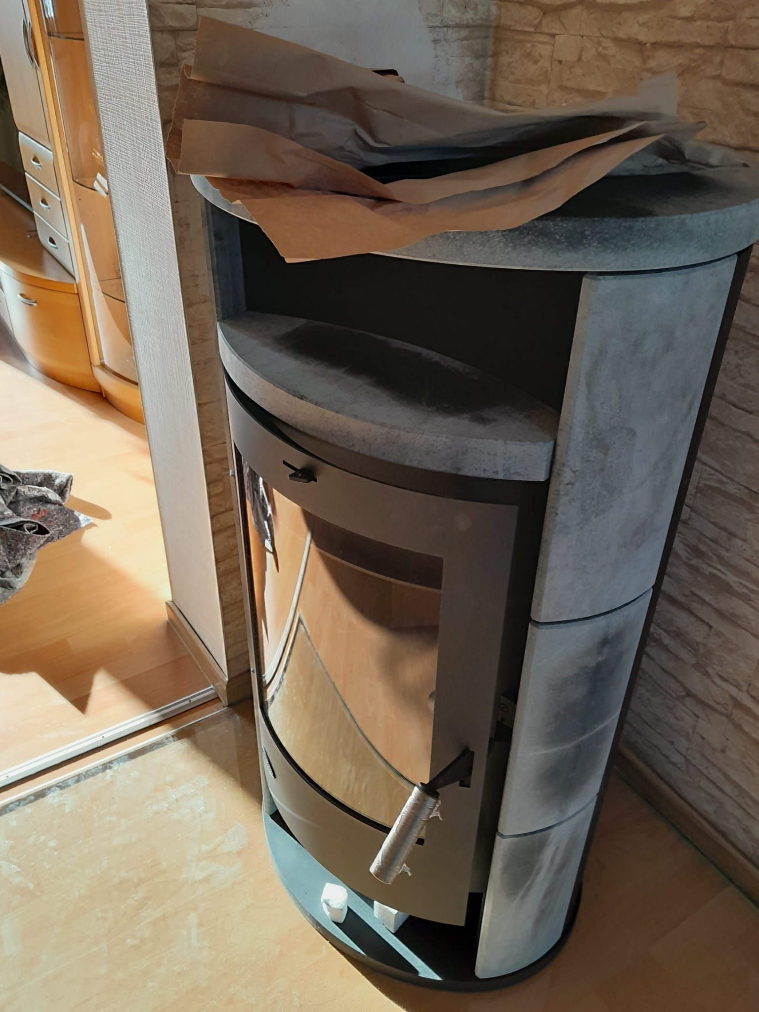 Grauer Steinofen mit geschwungener Front und Glastür, auf Holzboden stehend.