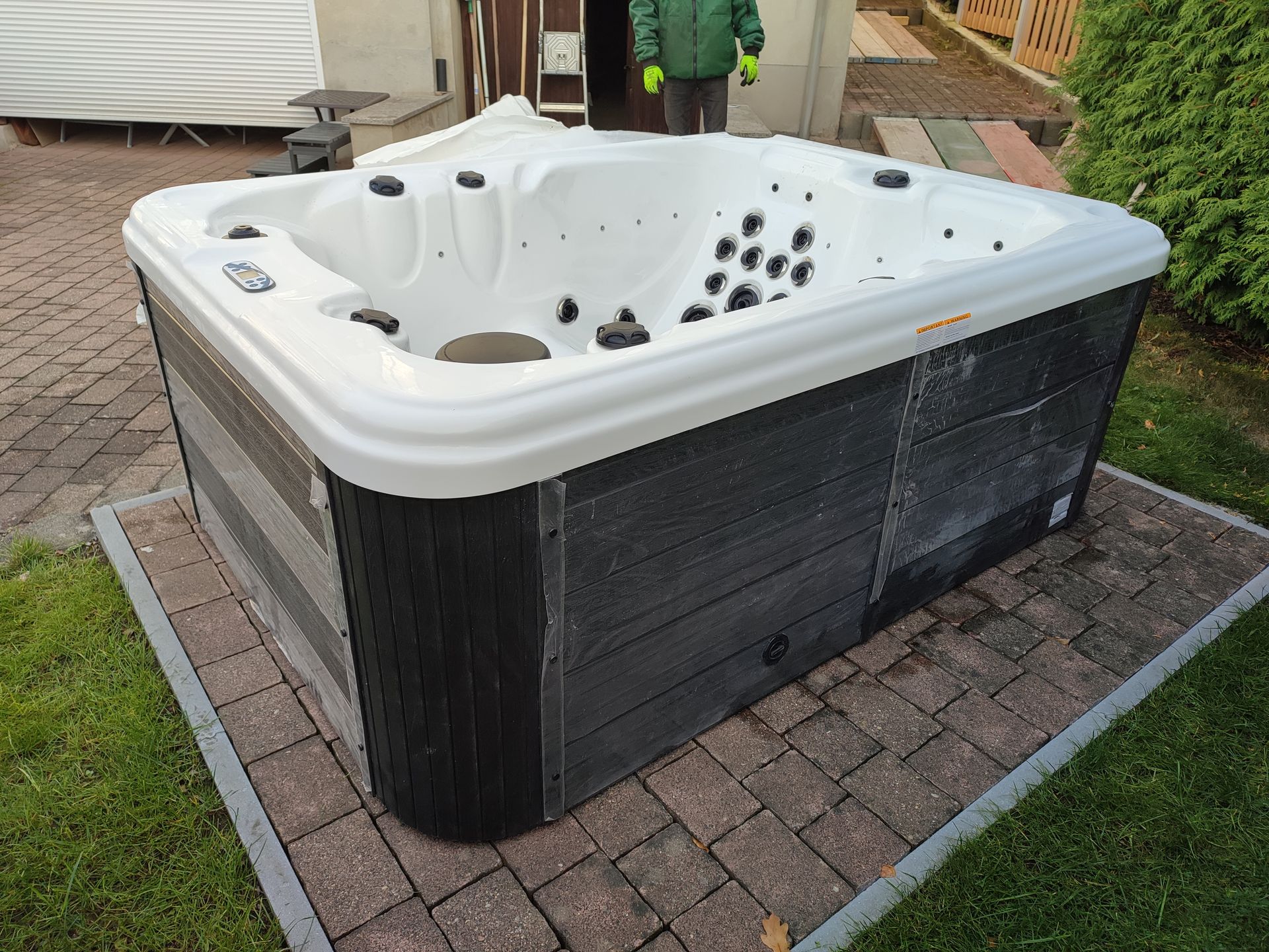 Whirlpool mit grauer und schwarzer Verkleidung auf einer mit Ziegeln bemauerten Terrasse, umgeben von Rasen.