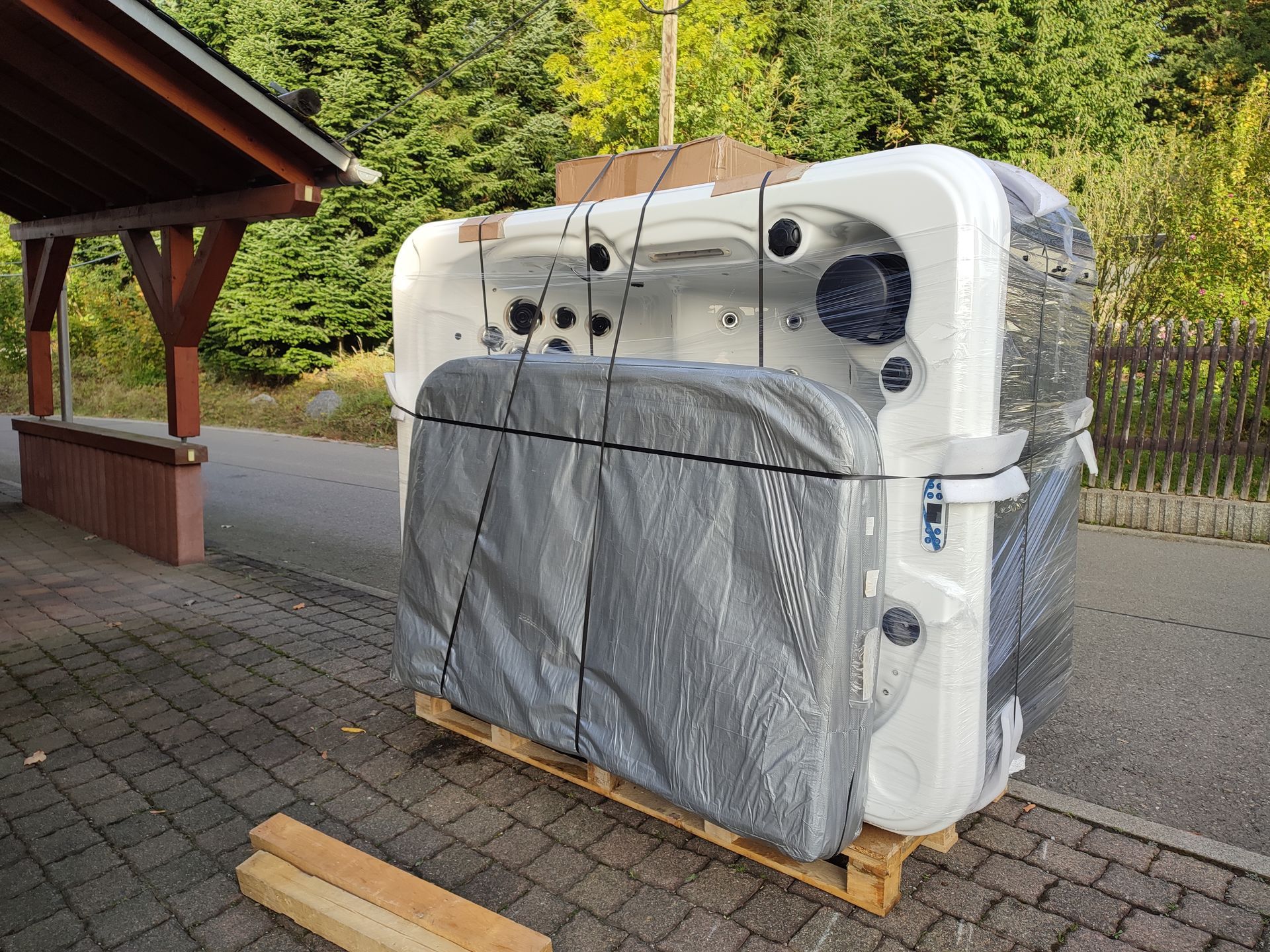 Ein verpackter weißer Whirlpool auf einer Holzpalette, in graue Plastikfolie eingehüllt, in der Nähe eines Holzgebäudes und Bäumen.