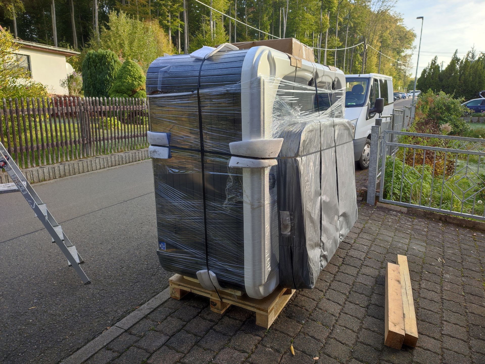 Ein eingewickelter Whirlpool auf einer Palette, abgestellt auf einer gepflasterten Straße neben einem Zaun.