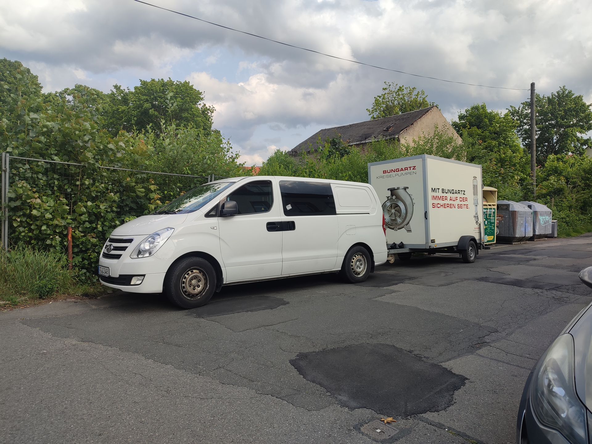 Ein weißer Lieferwagen mit Anhänger parkt auf einer Straße mit Mülltonnen. Im Hintergrund sind Bäume und ein Gebäude zu sehen.