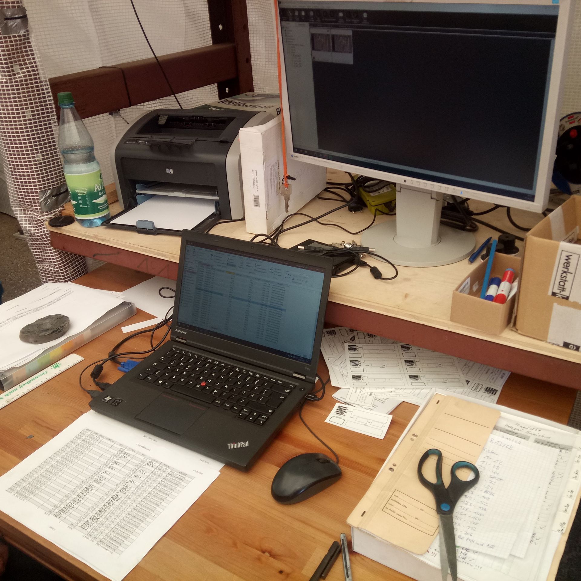 Laptop und Desktop-Monitor auf einem Schreibtisch, daneben Drucker, Papier, Schere und eine Wasserflasche.