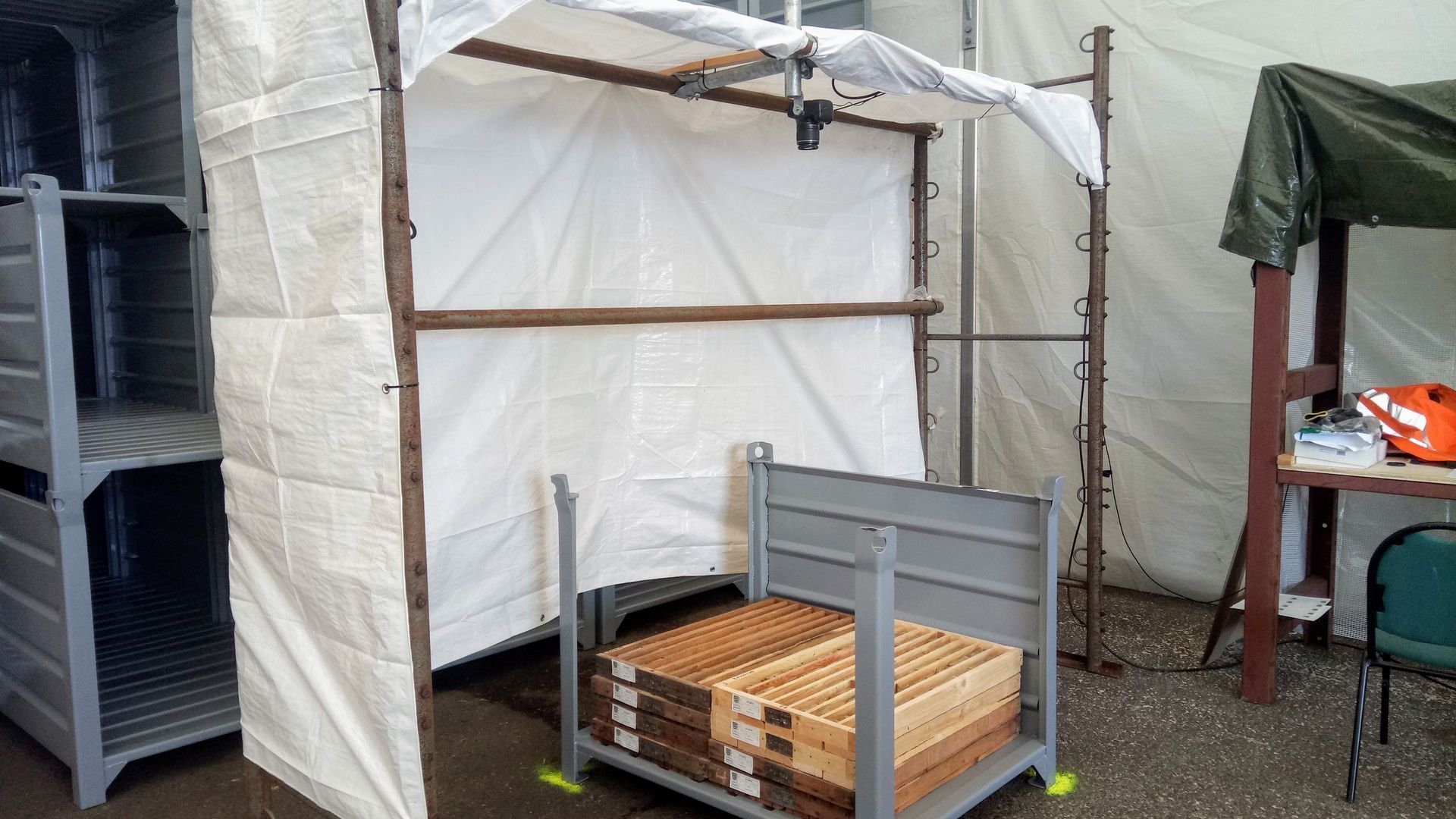 Behelfsmäßige Unterkunft mit Metallrahmen und weißer Plane über einem Stapel Holzkisten.