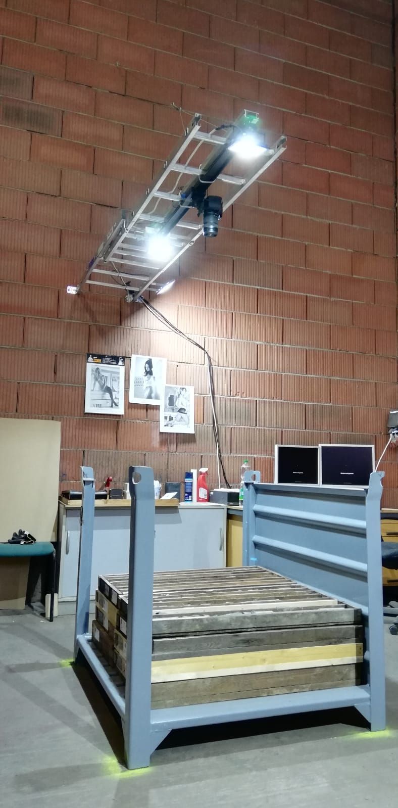 Ein Bettgestell mit einer Holzmatratze unter einer montierten Lichtanlage an einer Backsteinwand.