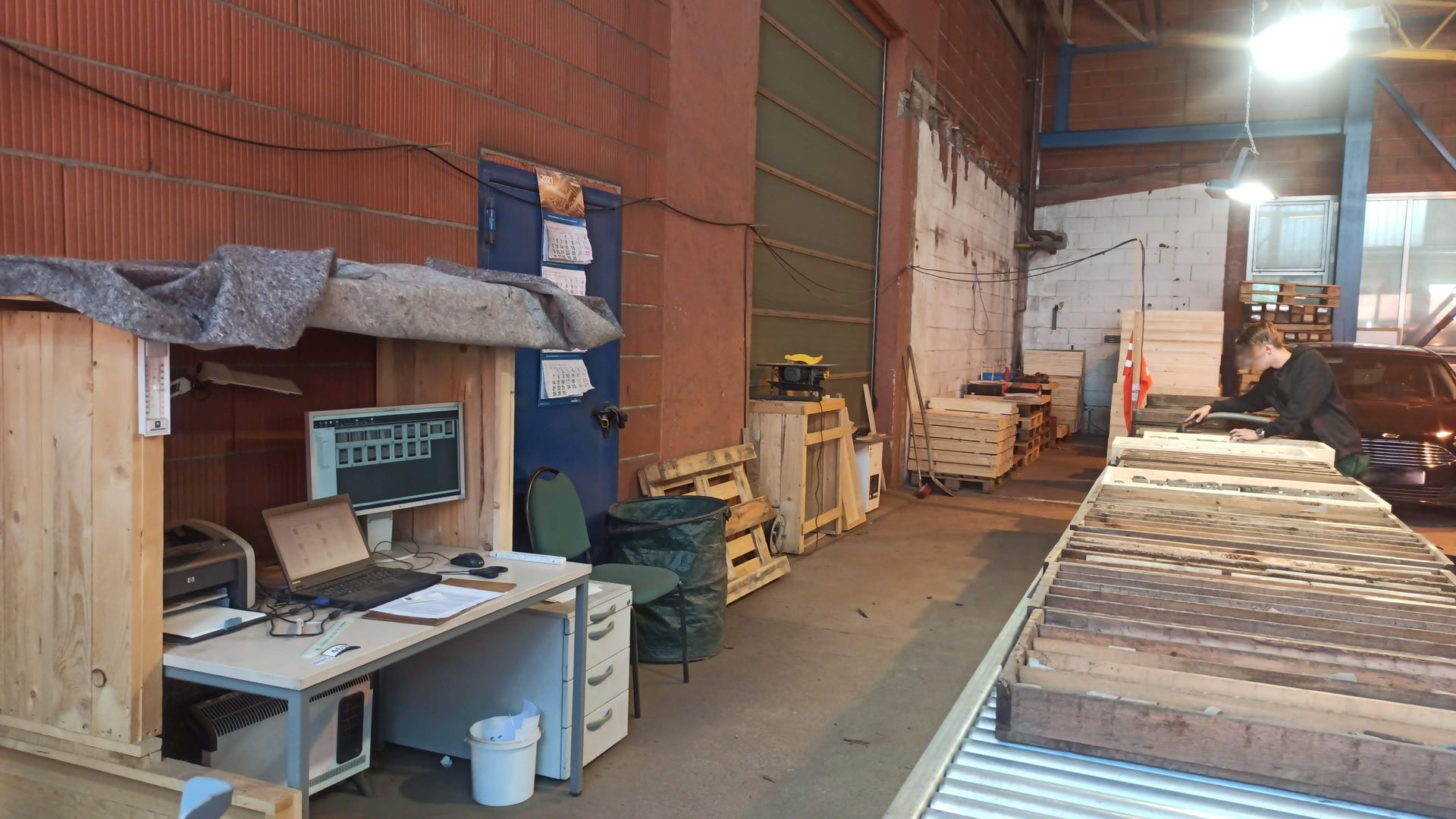 Arbeitsplatz im Innenbereich mit Schreibtisch, Computer, Drucker und einer Person, die an einem langen Tisch arbeitet; die Wände sind rot, grün und weiß.