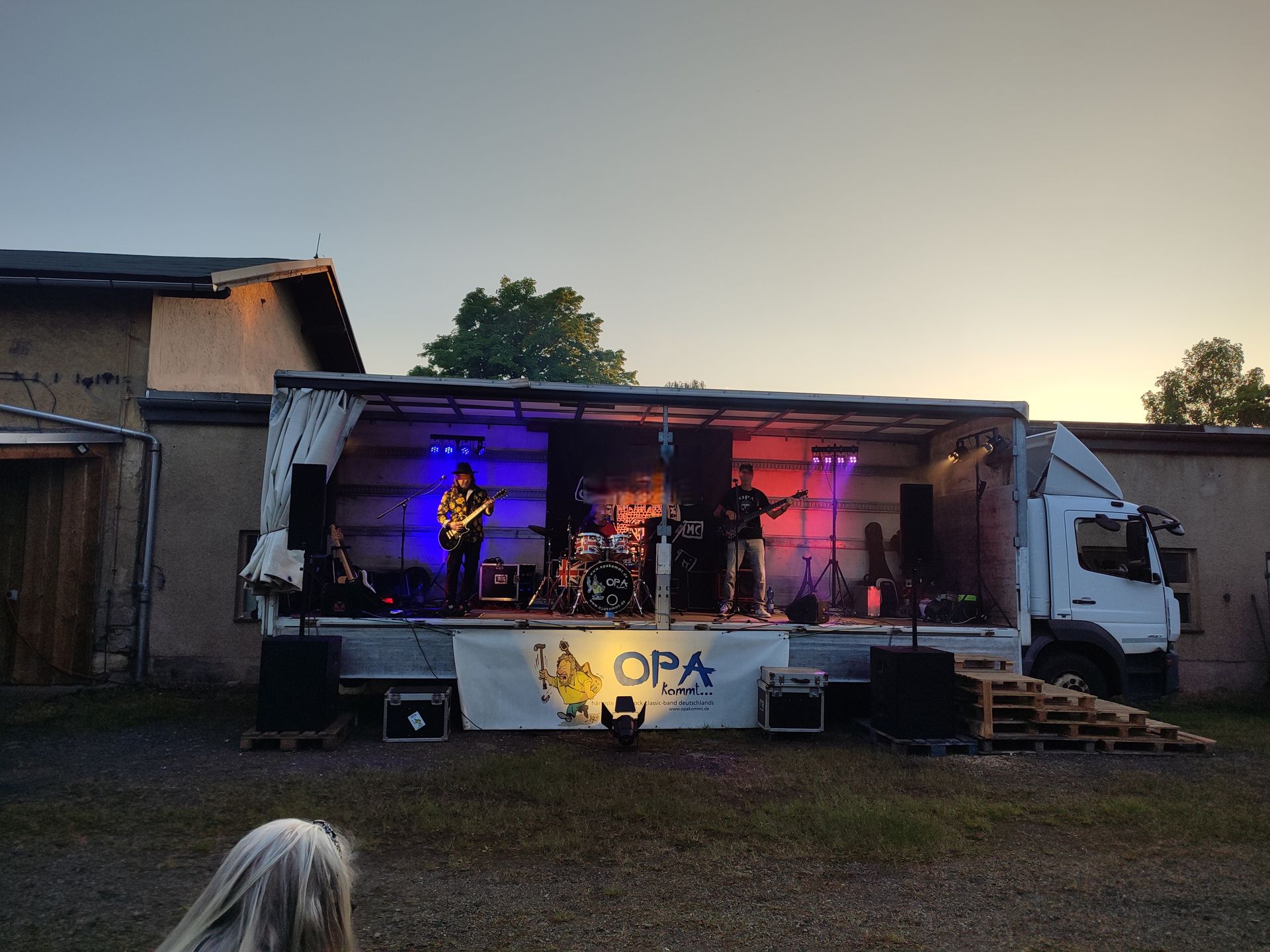 Eine Band spielt in der Abenddämmerung auf einer LKW-Bühne im Freien; die Bühne ist blau und rot beleuchtet.
