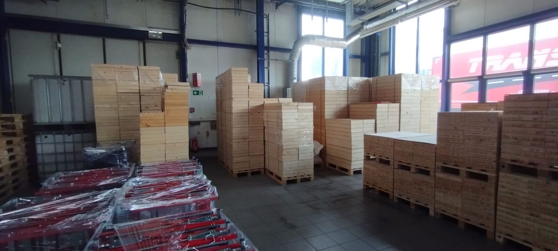 Innenansicht einer Lagerhalle mit gestapelten Holzkisten und Paletten. Einige Gegenstände sind in Plastikfolie eingewickelt; rechts befinden sich Fenster.