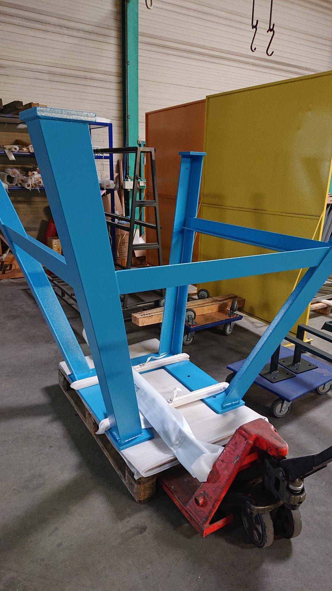 Blauer Metallrahmen auf einer Palette, die auf einem roten Hubwagen in einer Werkstatt ruht.