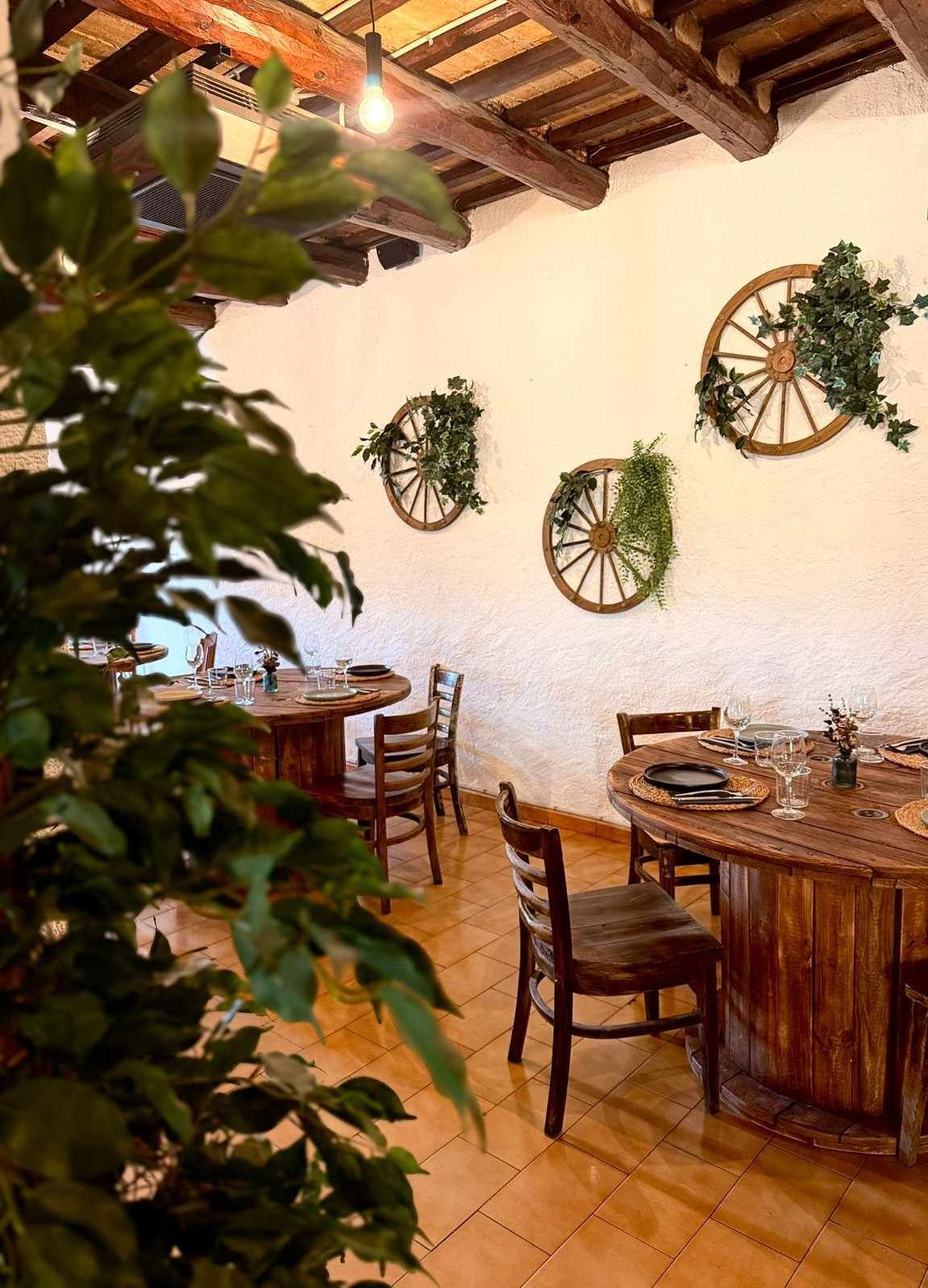Patio rústico de restaurante con mesas y sillas de madera, plantas colgantes y un mural decorativo con forma de rueda.