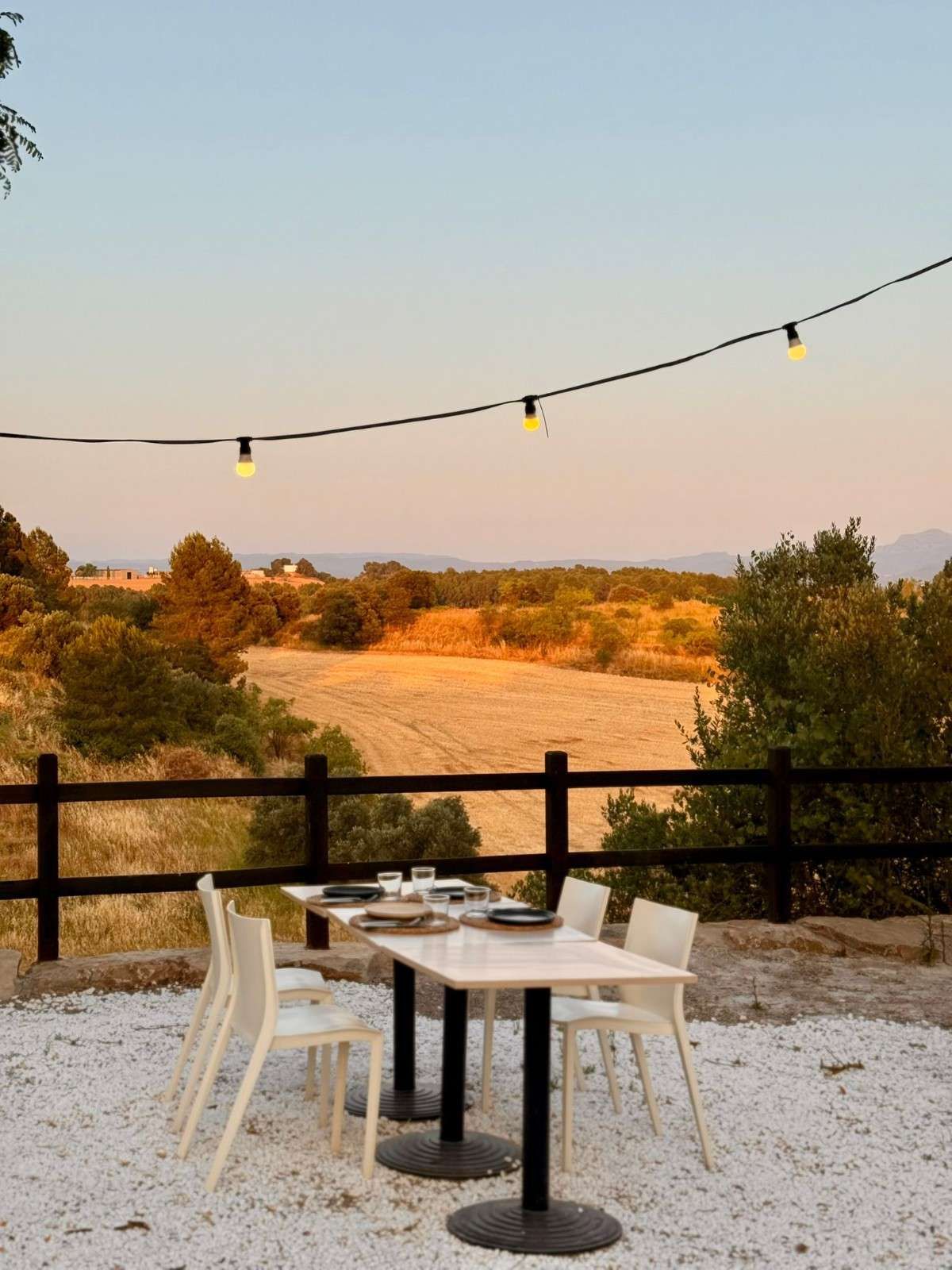 Mesa de patio exterior con sillas blancas, guirnaldas de luces y vistas a la puesta de sol sobre los campos.