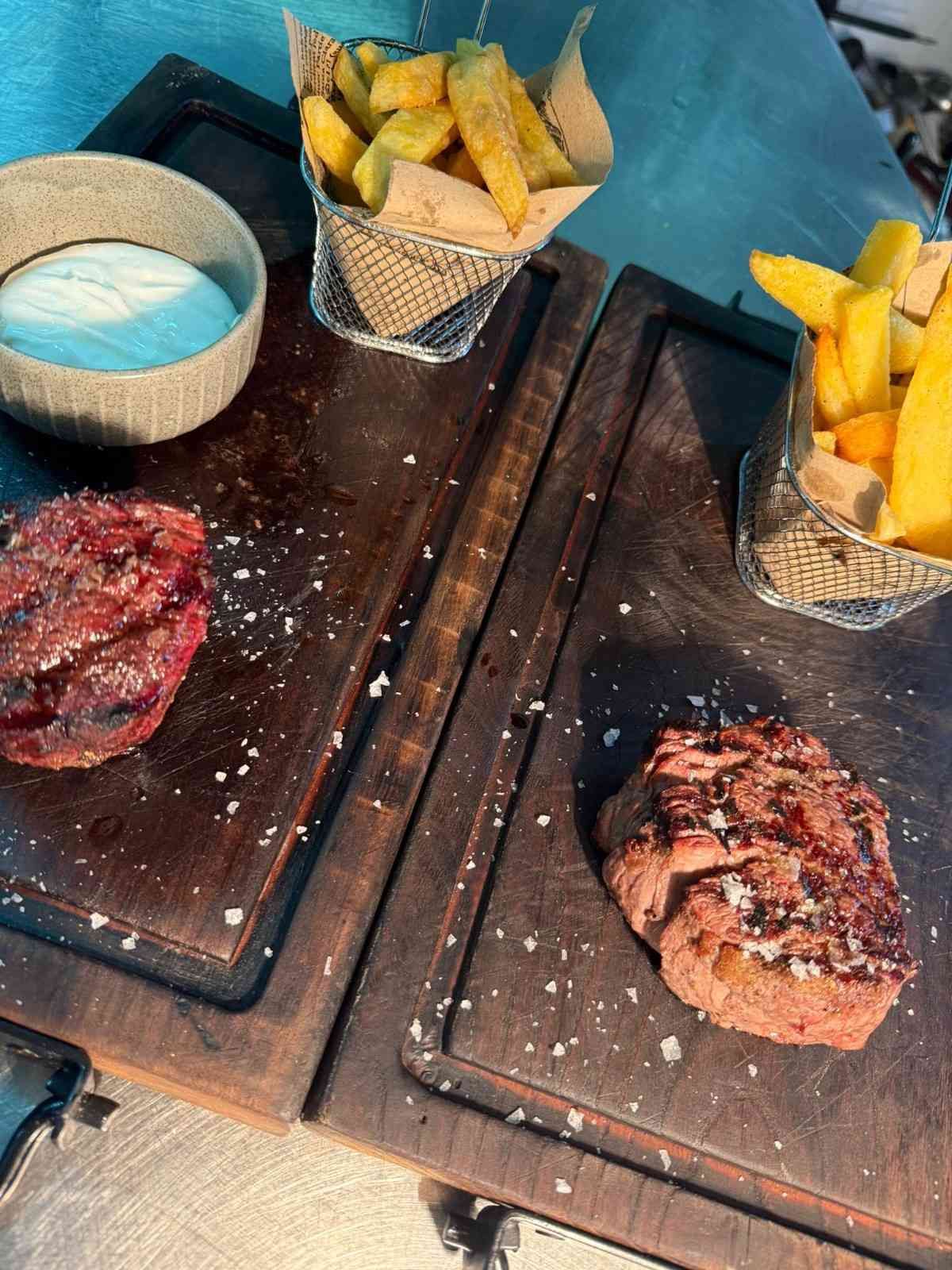 Dos filetes a la parrilla sobre tablas de madera con patatas gajo y vasitos de salsa para mojar sobre una mesa.