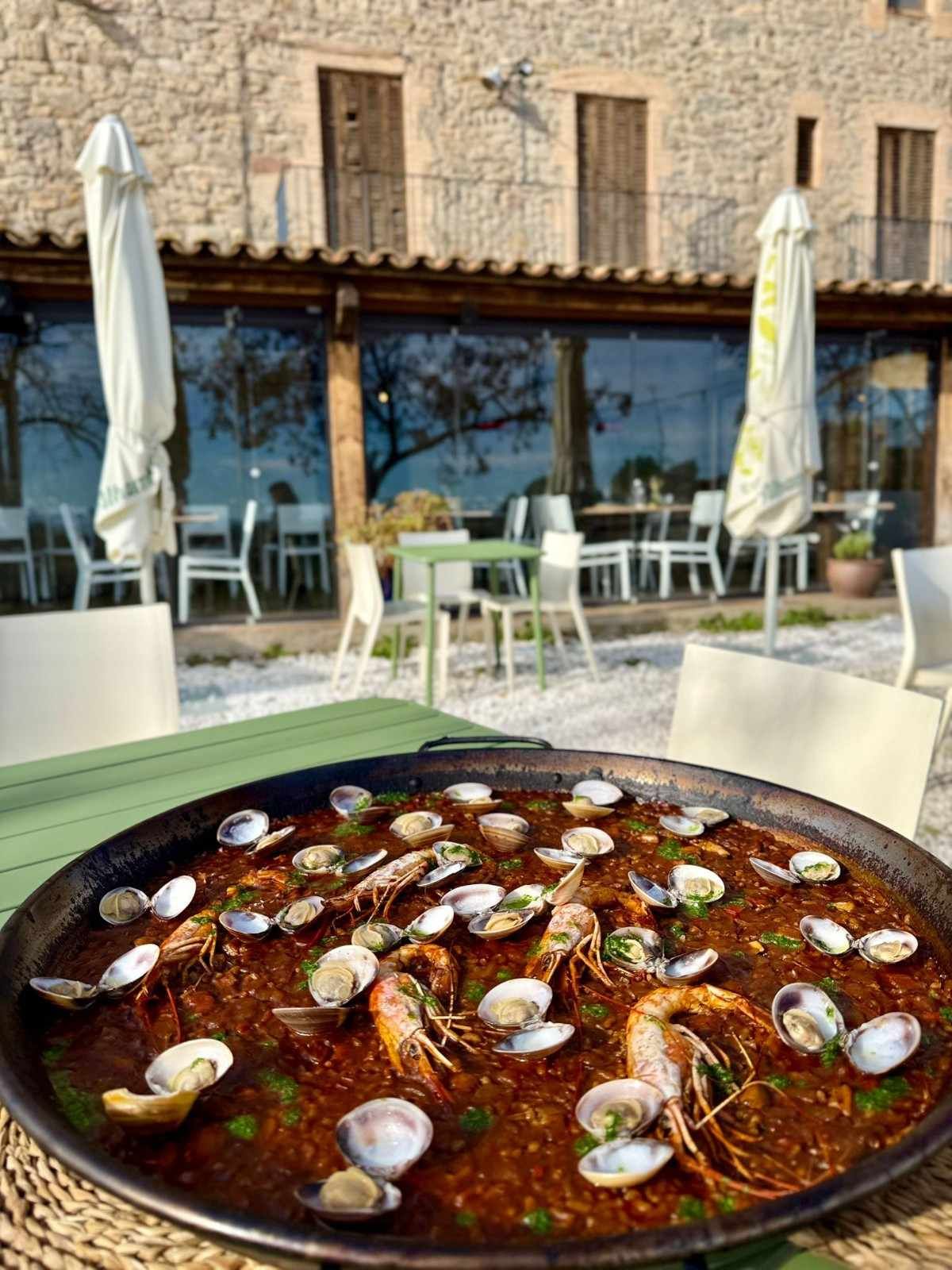 Mesa de restaurante al aire libre con una gran paella de mariscos, sillas blancas y sombrillas cerradas