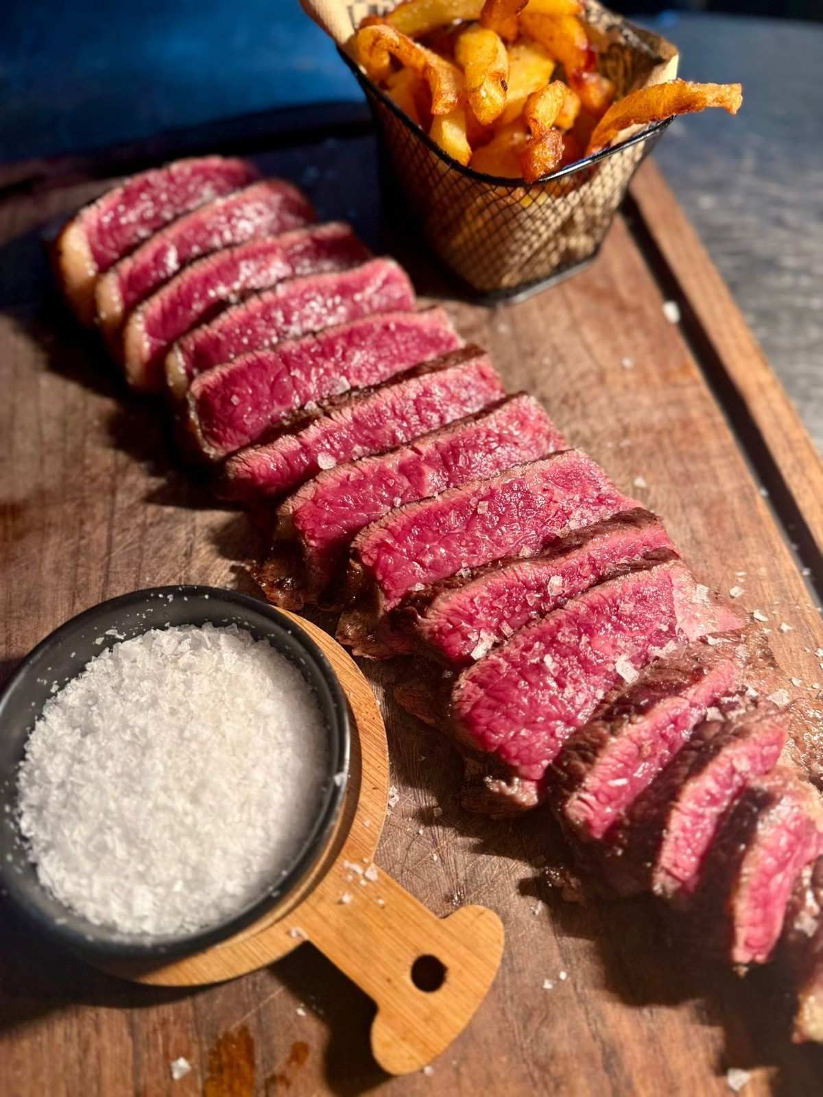 Filete de ternera poco hecho en rodajas con patatas fritas y un cuenco de sal gruesa sobre una tabla de madera.