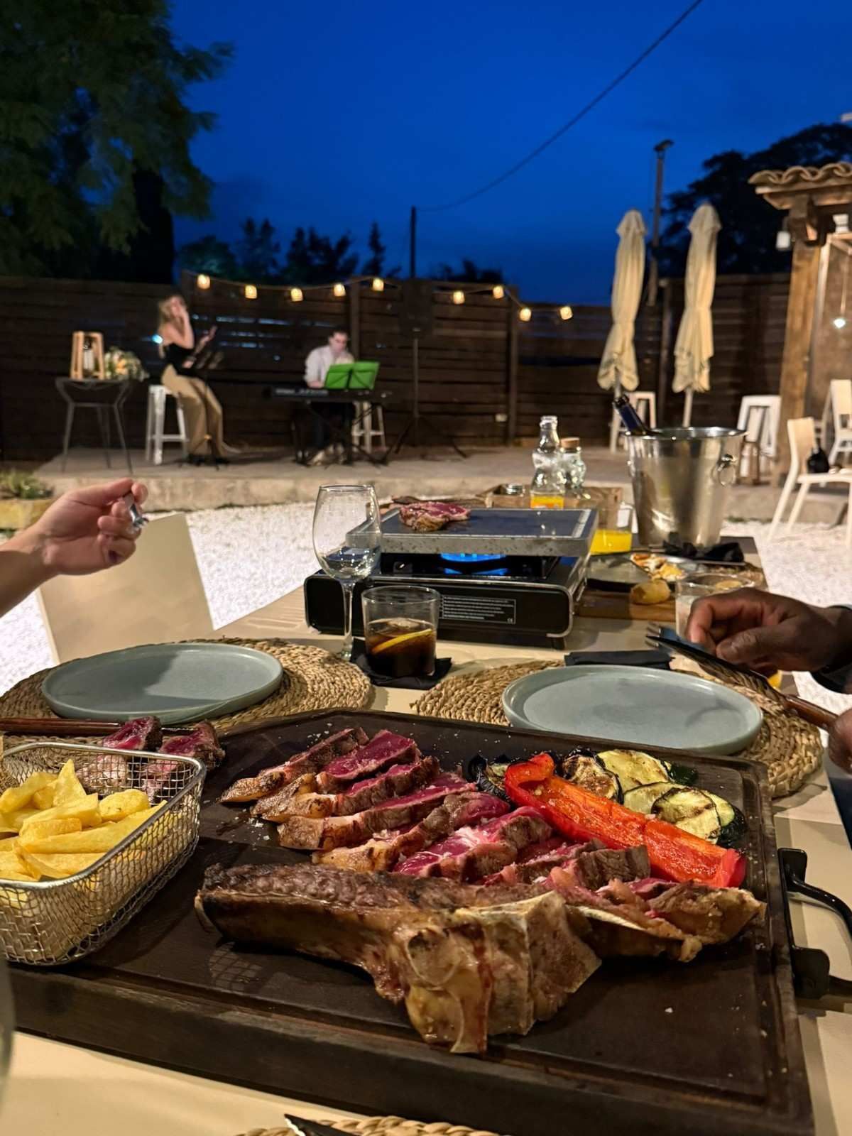 Cena al aire libre con carne a la parrilla, papas fritas y bebidas al atardecer en un patio.