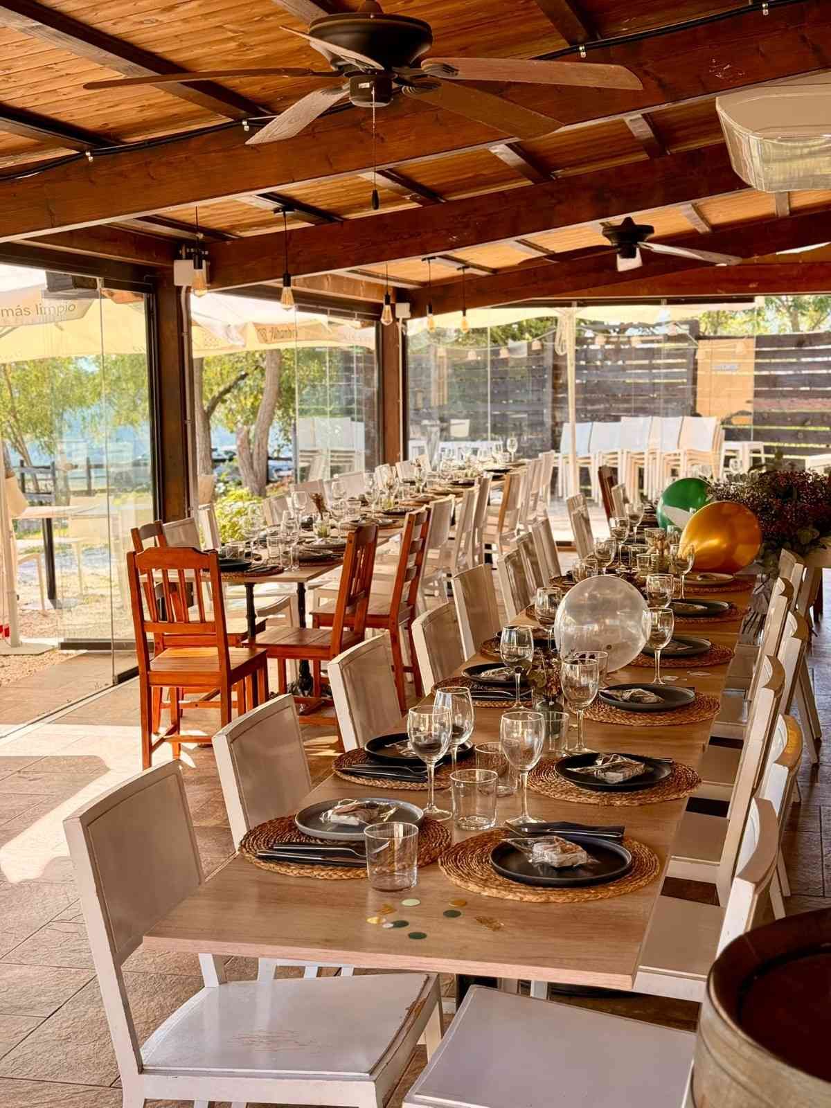 Mesa larga con platos y cristalería bajo un techo de madera en el patio exterior.