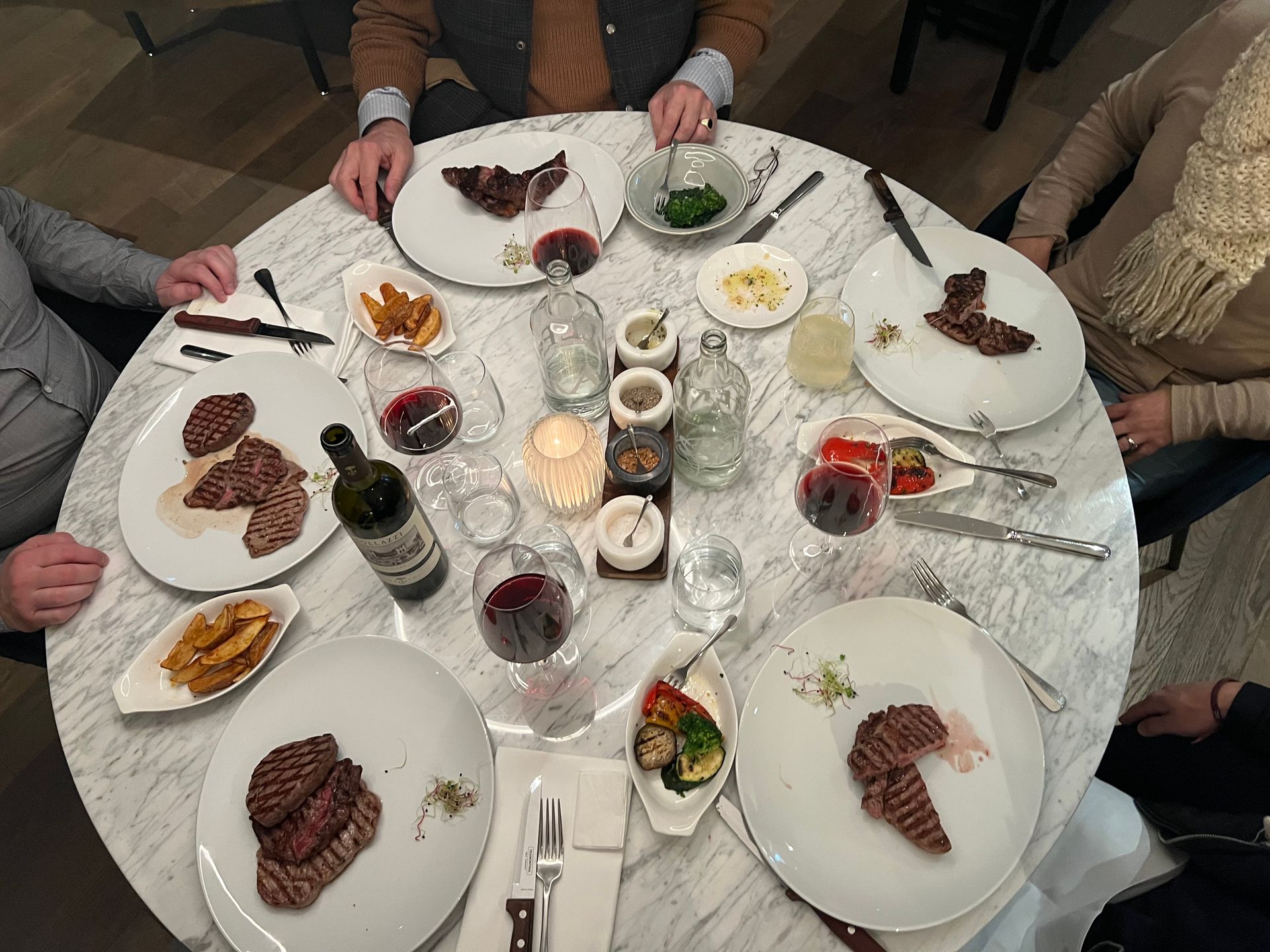 Mehrere Teller mit Speisen und Getränken auf einem dunklen Tisch: Steak, Lammkoteletts, Hühnchen, Kroketten, Bier und Wein.