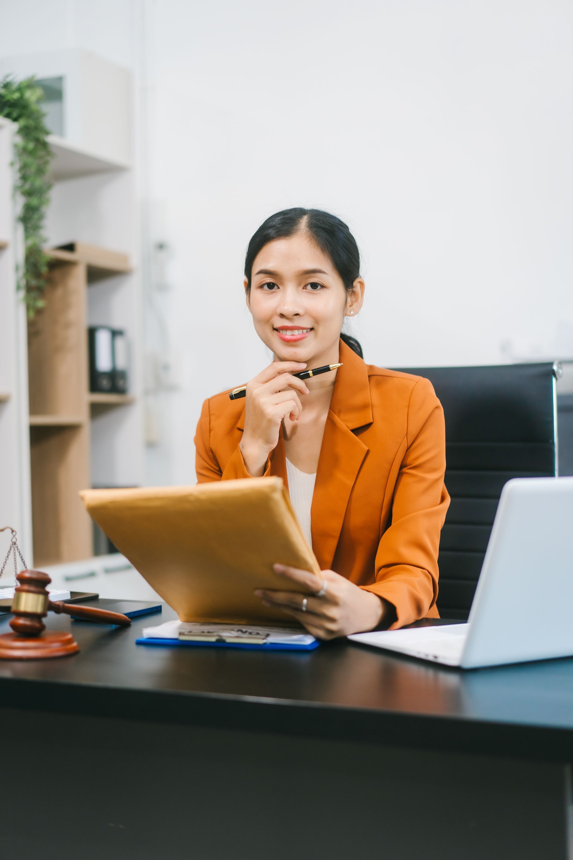 Femme en blazer orange, tenant un dossier, souriante à son bureau avec un ordinateur portable et un maillet.
