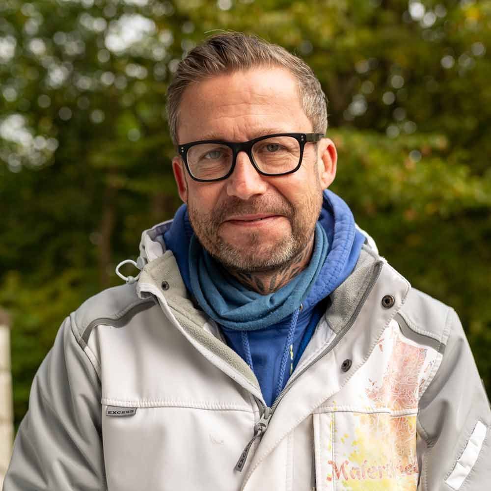 Ein Mann mit Brille und Jacke steht vor Bäumen.