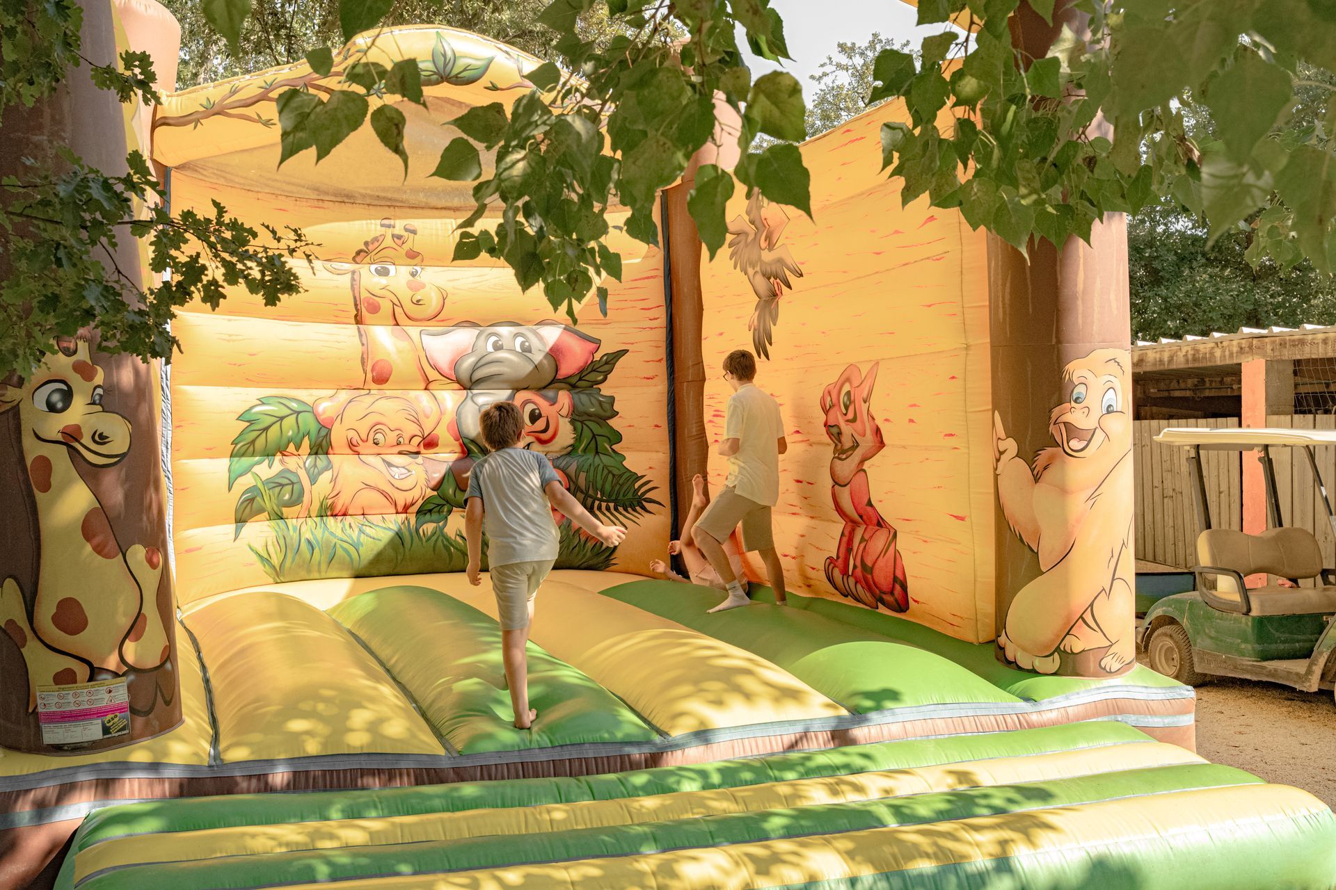 Des enfants jouent dans un château gonflable à thème animalier. Couleurs jaunes et vertes, en extérieur.