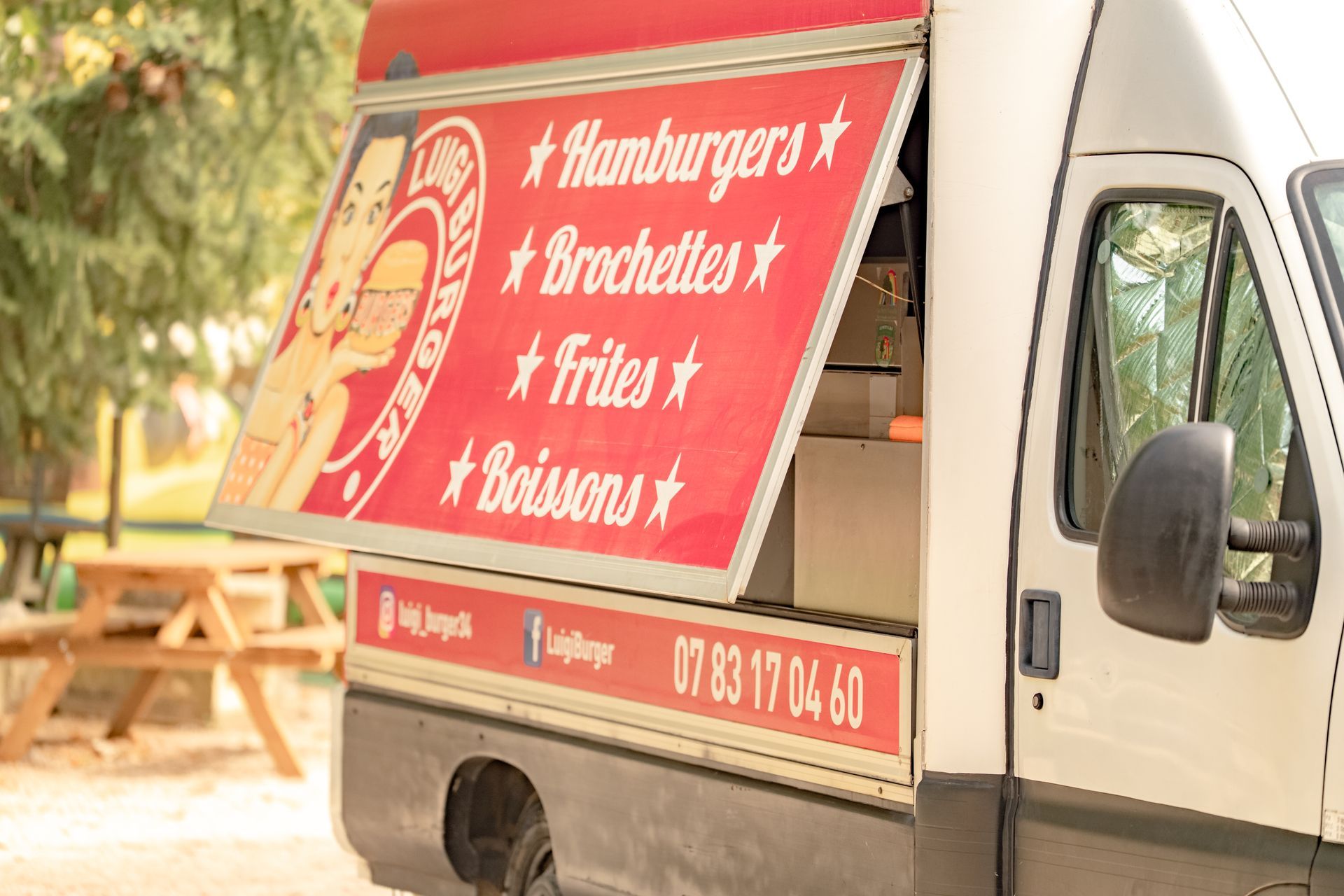Camionnette rouge et blanche proposant un menu composé de hamburgers, brochettes, frites et boissons. Fenêtre ouverte, en terrasse.