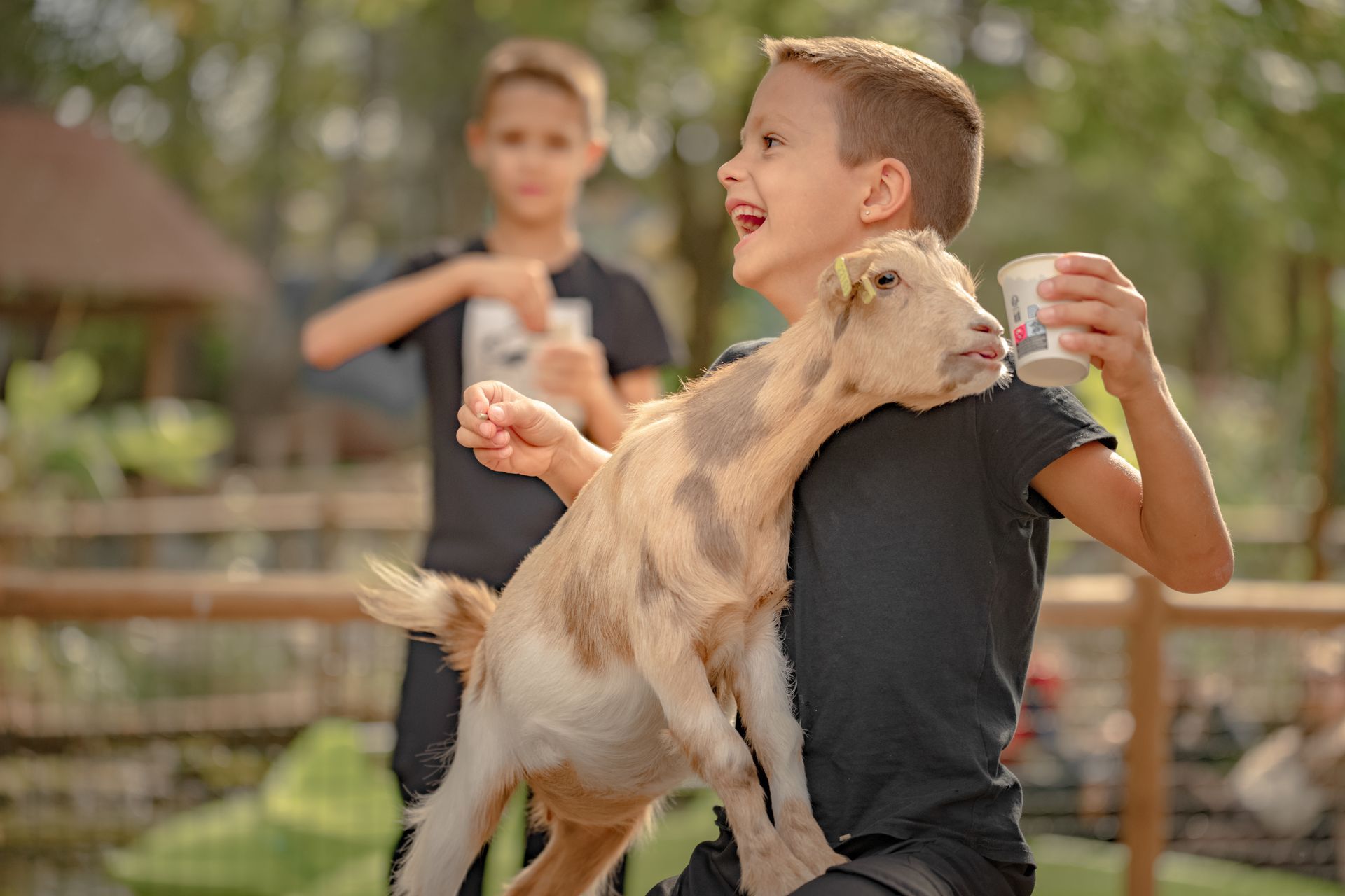 Un garçon tenant une petite chèvre, souriant avec une tasse ; un autre garçon à l'arrière-plan.
